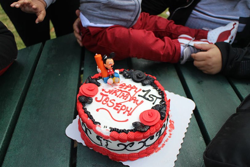 Birthday cake with Mickey Mouse figurine, red and black decorations, and red roses, celebrating Joseph's first birthday, on a green picnic table