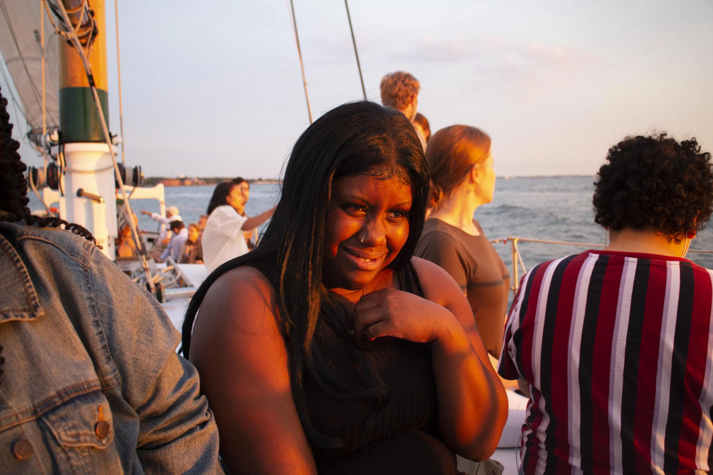 A group of people on a boat enjoying a sunset, with a woman in the foreground smiling and looking at the camera, and others looking at the water and scenery.