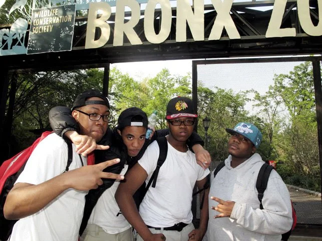 Four young boys standing together outdoors at the Bronx Zoo, with a sign for the Wildlife Conservation Society above them. They are wearing casual clothes and backpacks, and the background shows green trees and a fence.