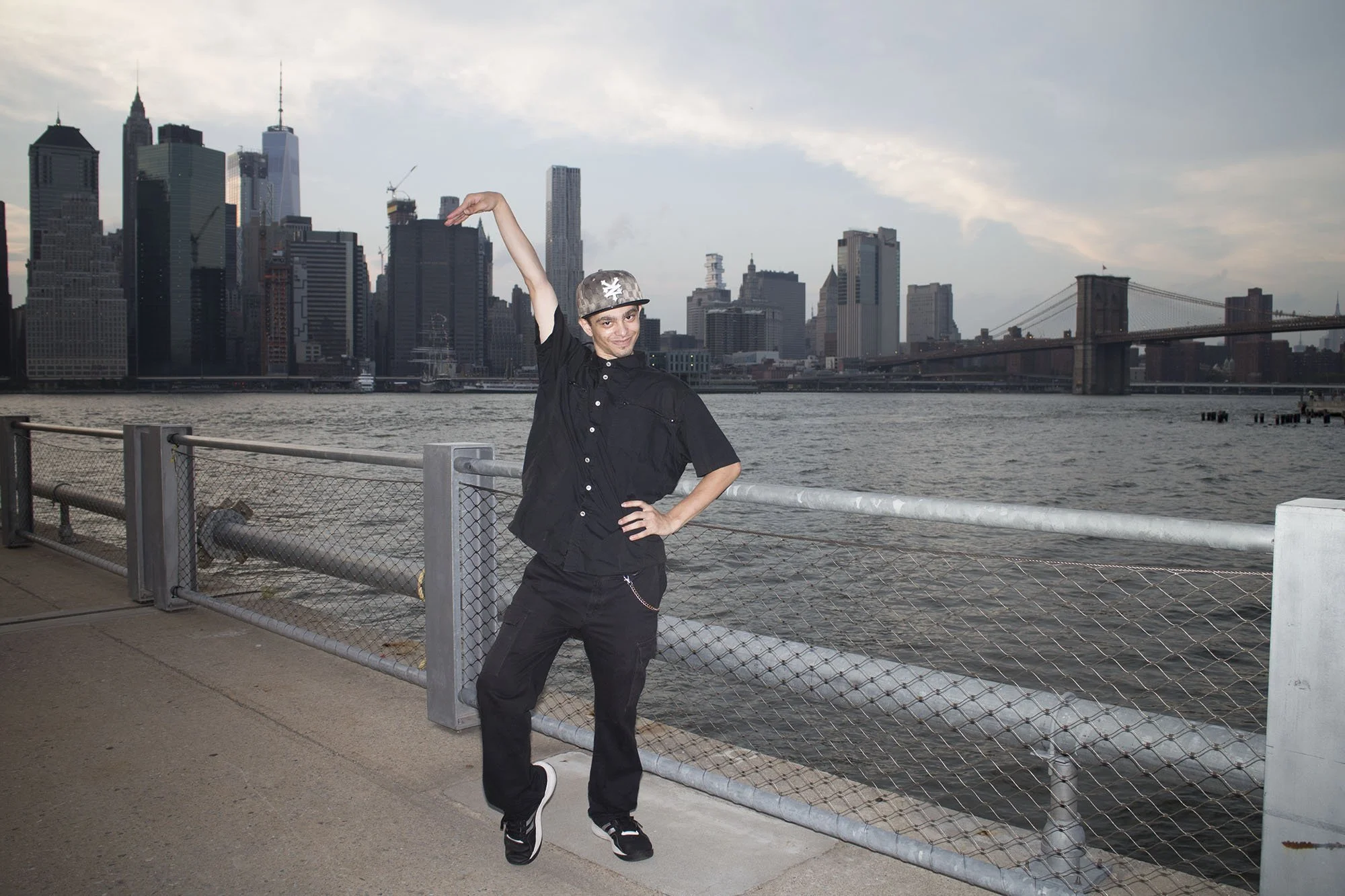 A person dressed in black with a chain hanging from their pants, wearing a cap, stands on a waterfront promenade with New York City skyline and Brooklyn Bridge in the background. The person is striking a pose with one arm raised and a smile.
