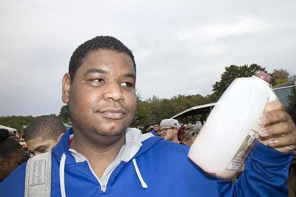 Young man in a blue jacket holding a large bottle or container outdoors, with a crowd and trees in the background.