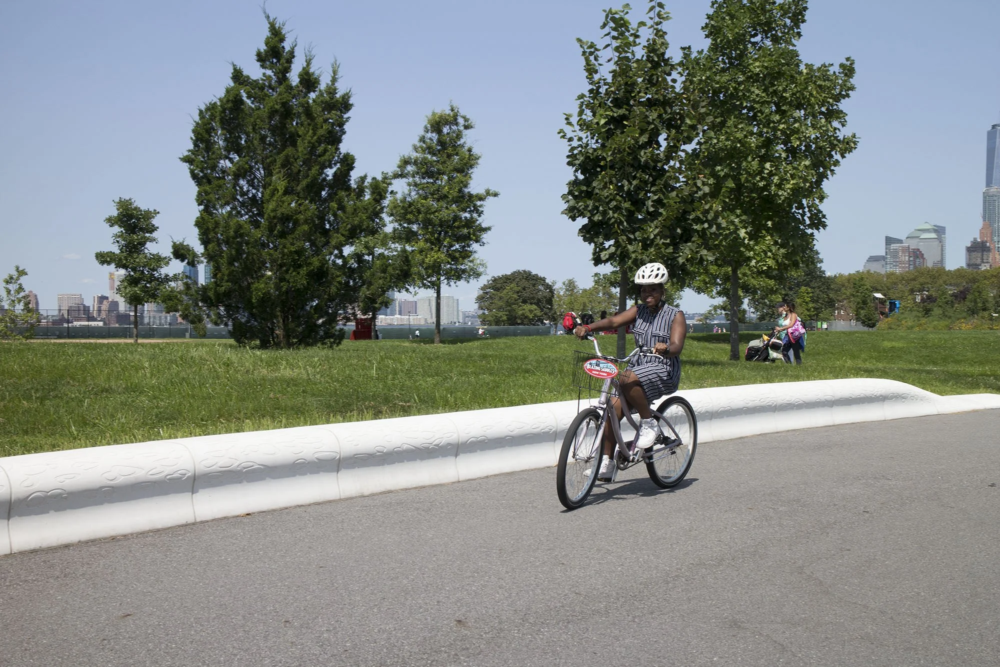 Woman riding a bicycle on a paved path in a park with trees and a city skyline in the background.