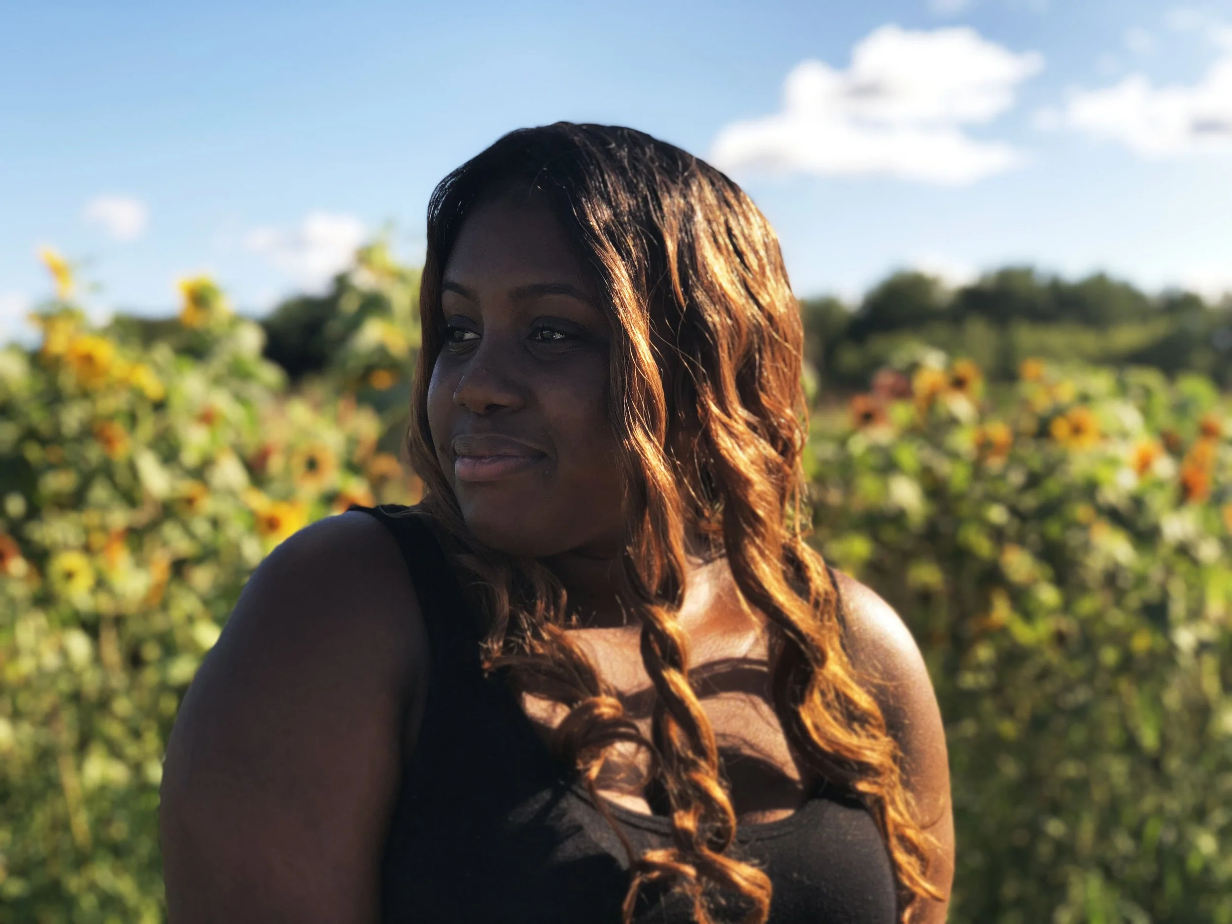 A woman with long, wavy hair standing outdoors in front of a field of yellow flowers under a blue sky with scattered clouds.
