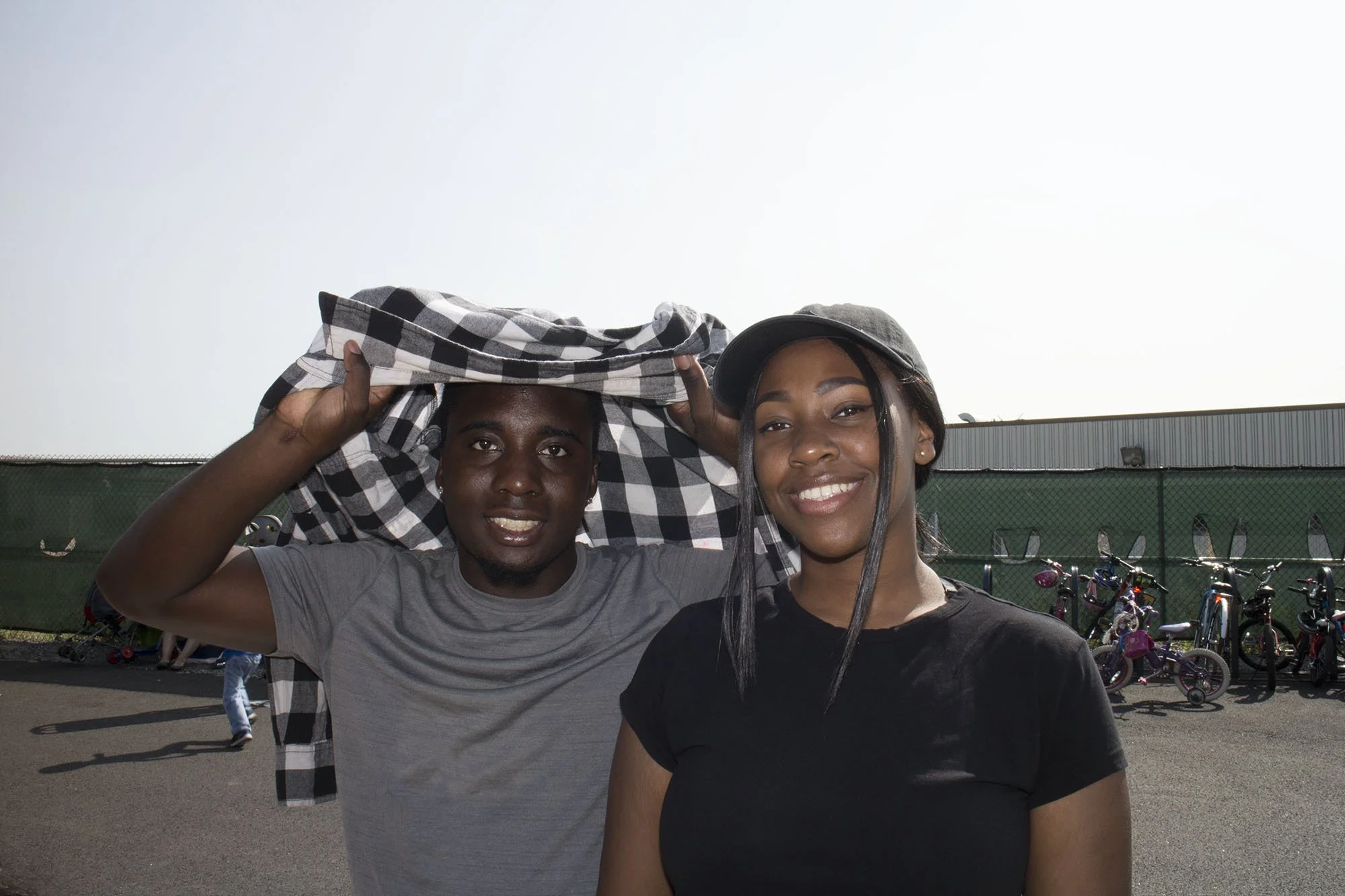 A smiling young man holding a checkered shirt above his head and a young woman standing beside him, both outdoors in a parking lot with bicycles and a fence in the background.