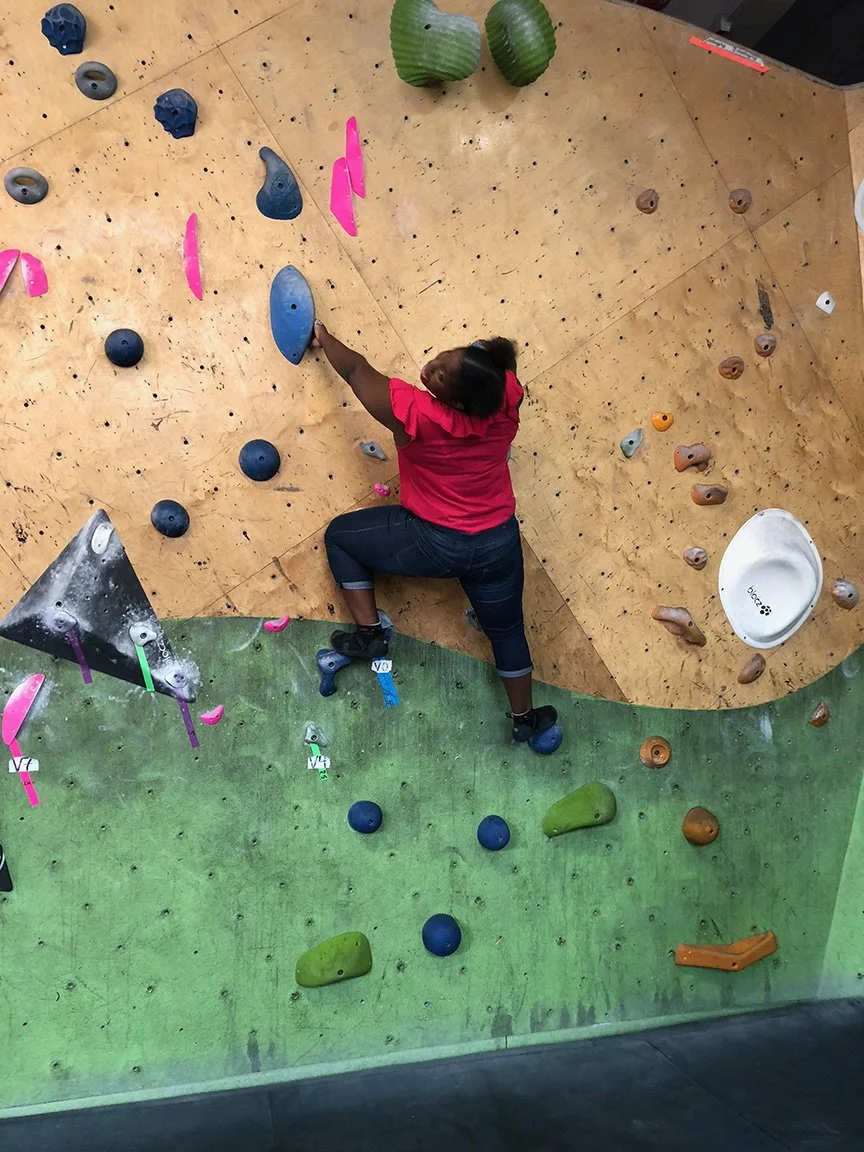 Person in pink shirt and dark jeans climbing a bouldering wall with various colored holds in an indoor rock climbing gym.