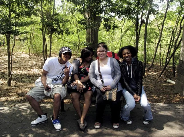Four women sitting on a park bench in a wooded area, smiling at the camera.