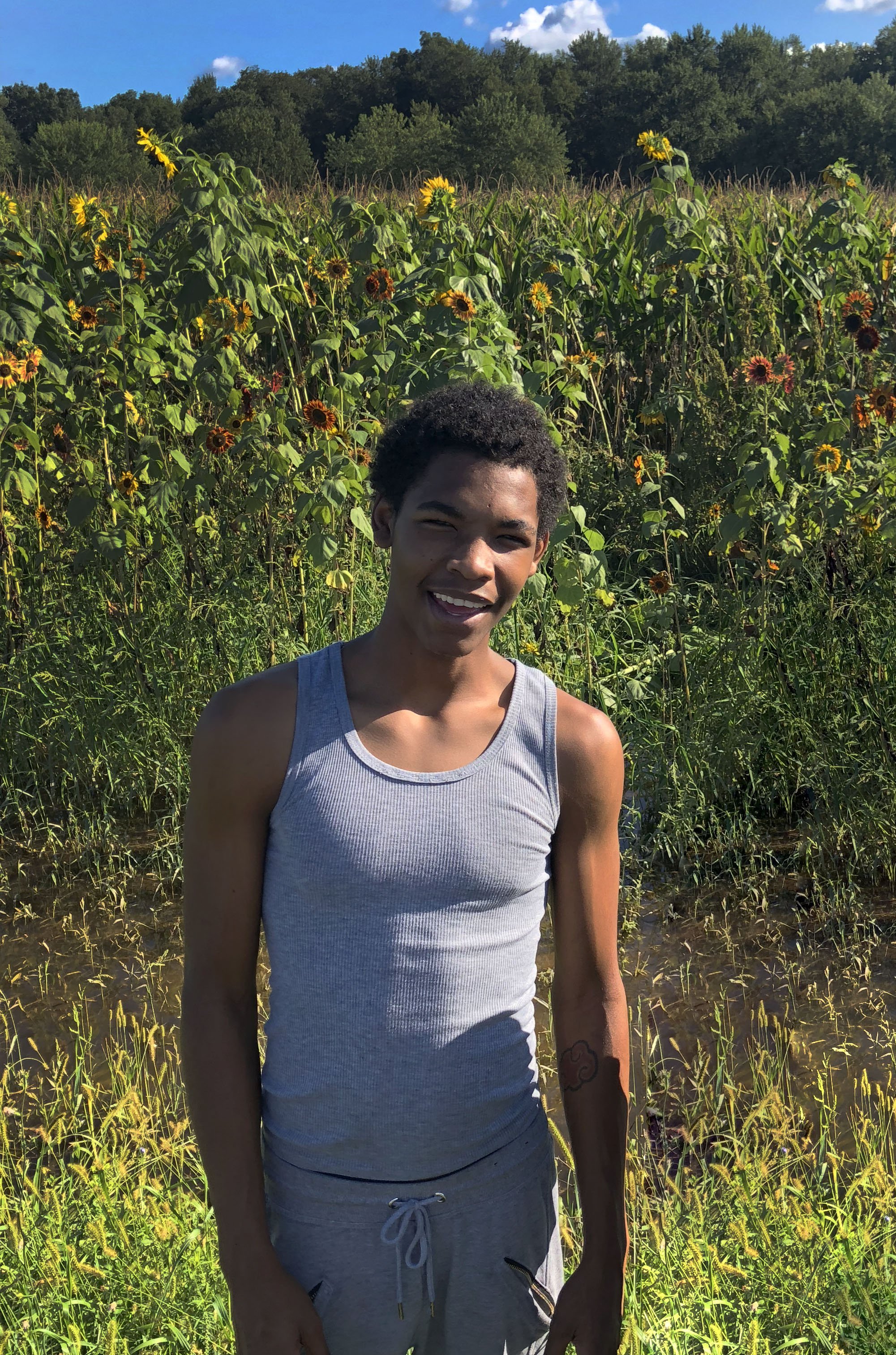 A young person smiling and standing in a field of tall sunflowers and other plants, with a background of trees and blue sky.