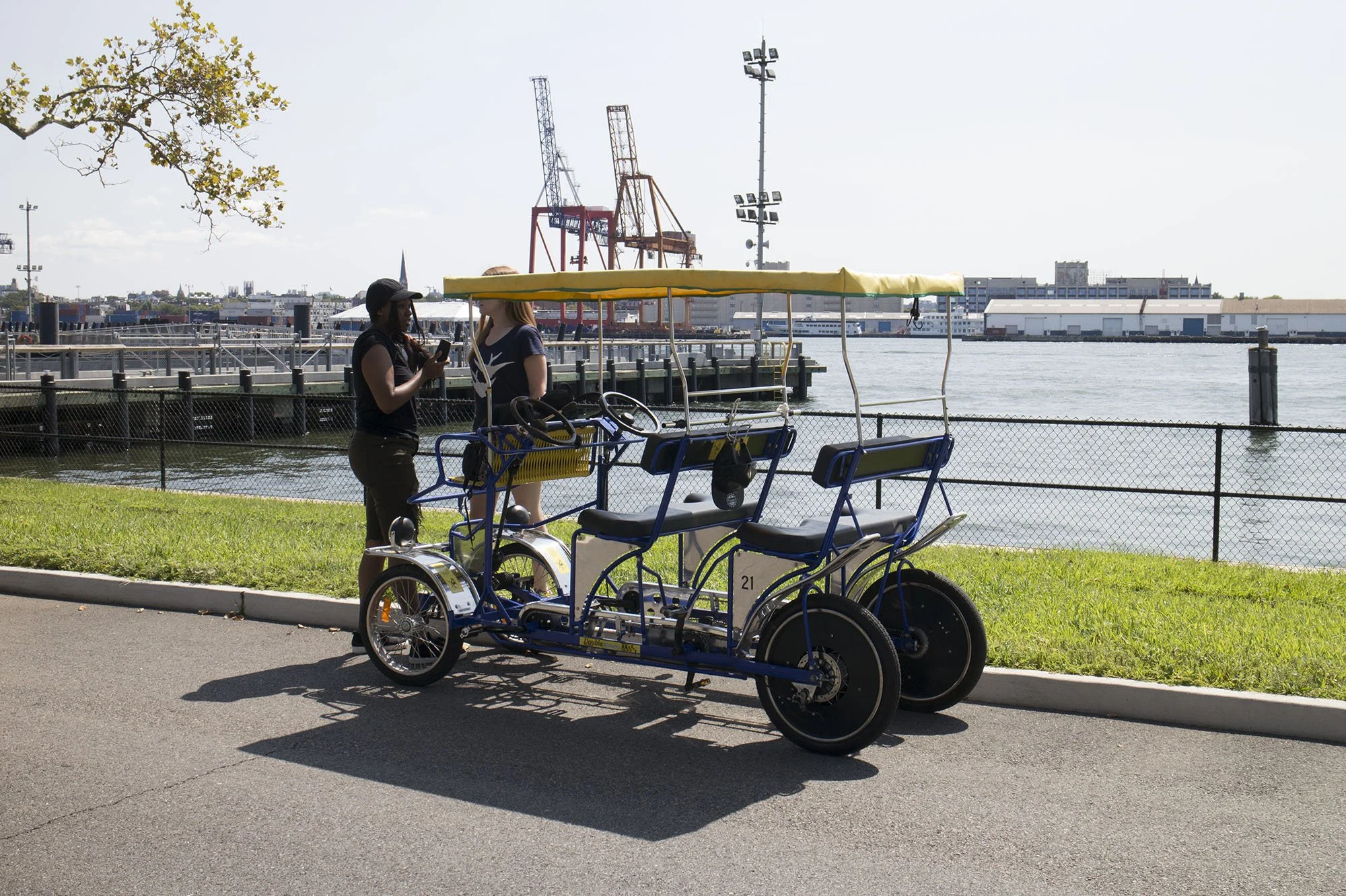 Two women standing next to a multi-passenger pedal bicycle built for four, parked by a waterfront with industrial cranes and buildings in the background.