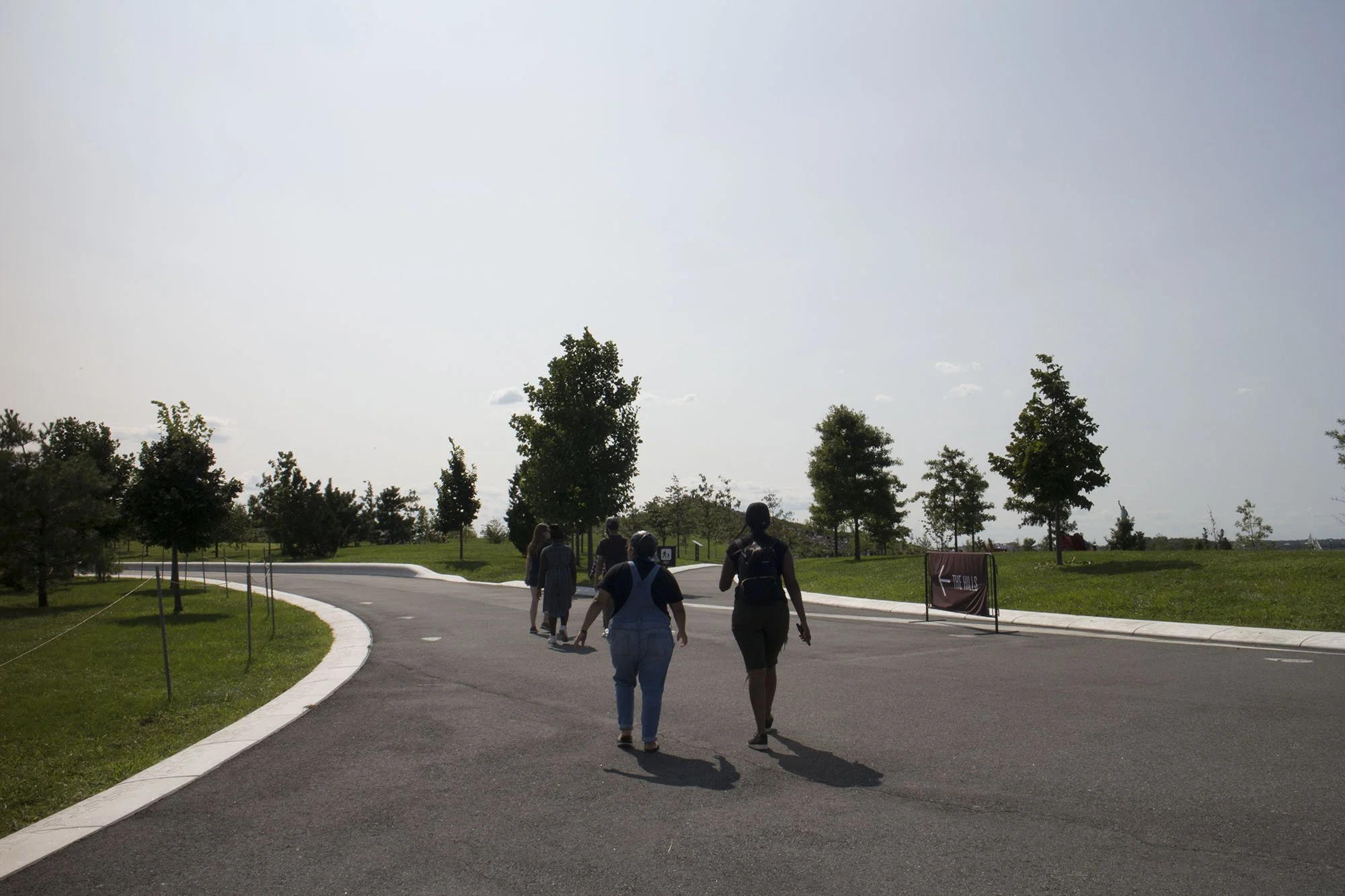 People walking on a paved path in a park with trees, grassy areas, and a partly cloudy sky.