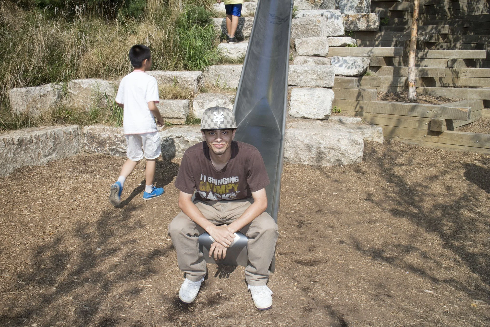 A young man sitting on a modern metal slide at a playground, smiling, with two children nearby, one walking away and another climbing stairs in the background.