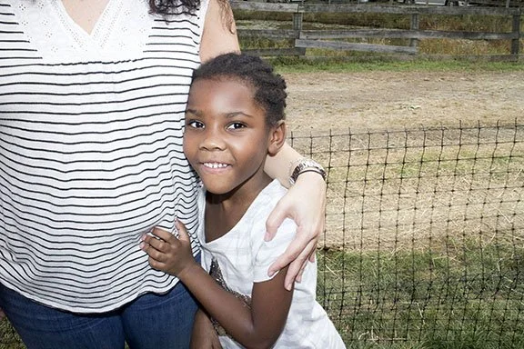A young girl with braided hair standing next to an adult, possibly a woman, outdoors near a wire fence and grassy area.