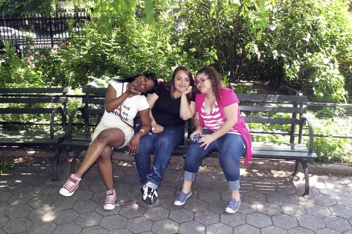Three girls sitting on a park bench under leafy trees, smiling and posing for the photo.