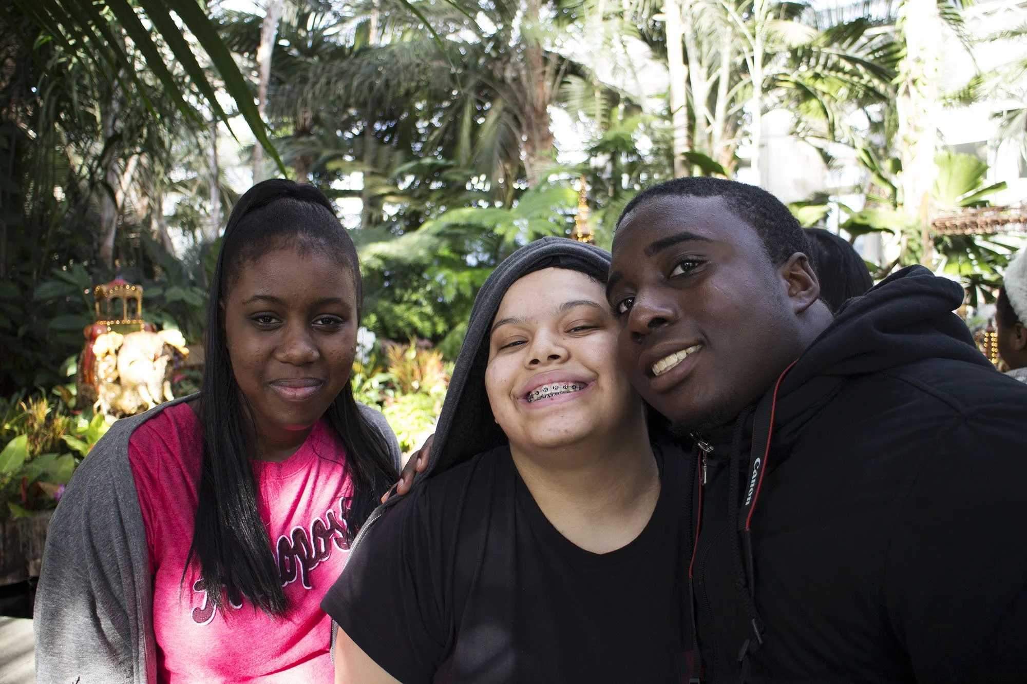 Three smiling young people taking a selfie in a lush indoor garden. The person in the middle has braces and a hoodie, the person on the left has long black hair and a pink shirt, and the person on the right has short hair and a black jacket.