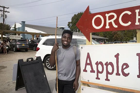 Young man smiling and holding a wooden stick next to a large sign that says 'Apple' and an arrow pointing toward an orchard.