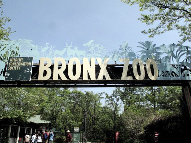 Entrance sign for Bronx Zoo with wildlife silhouettes and visitors walking nearby.