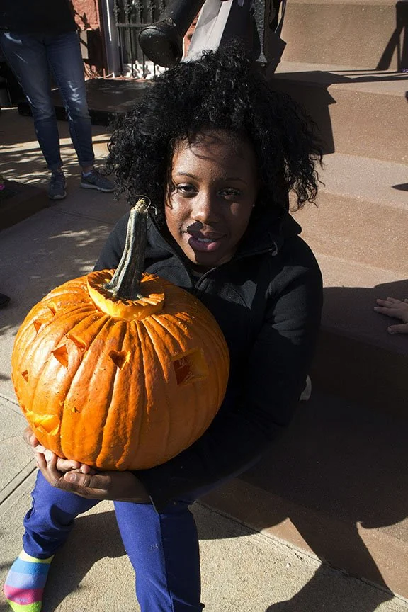 A young girl holding a large, carved pumpkin with a smile on her face. The pumpkin is bright orange with a green stem, and the girl is sitting outdoors on a step, wearing a black jacket and colorful striped socks.