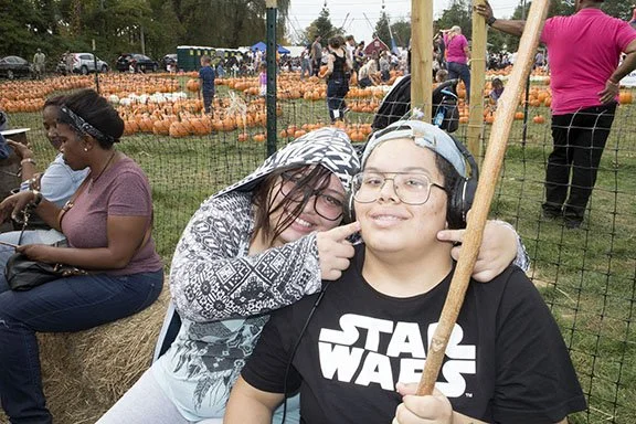 Two people, a girl and a boy with glasses, are at a pumpkin patch. The girl is touching the boy's ear and both are smiling. Other people and pumpkins are visible in the background.