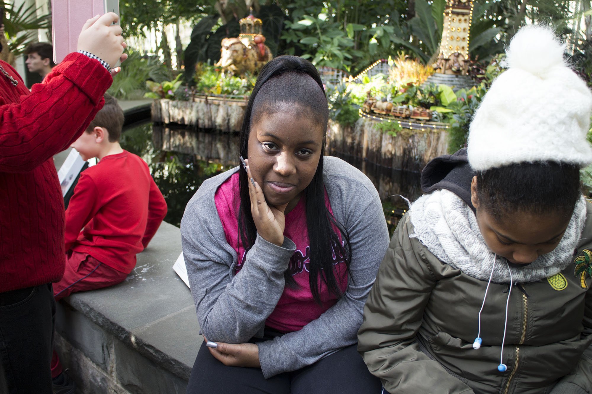 A woman with long black hair, wearing a pink shirt and gray hoodie, sitting next to a girl with a green jacket and white knit hat, in an indoor garden or conservatory with lush greenery and decorative elements in the background.
