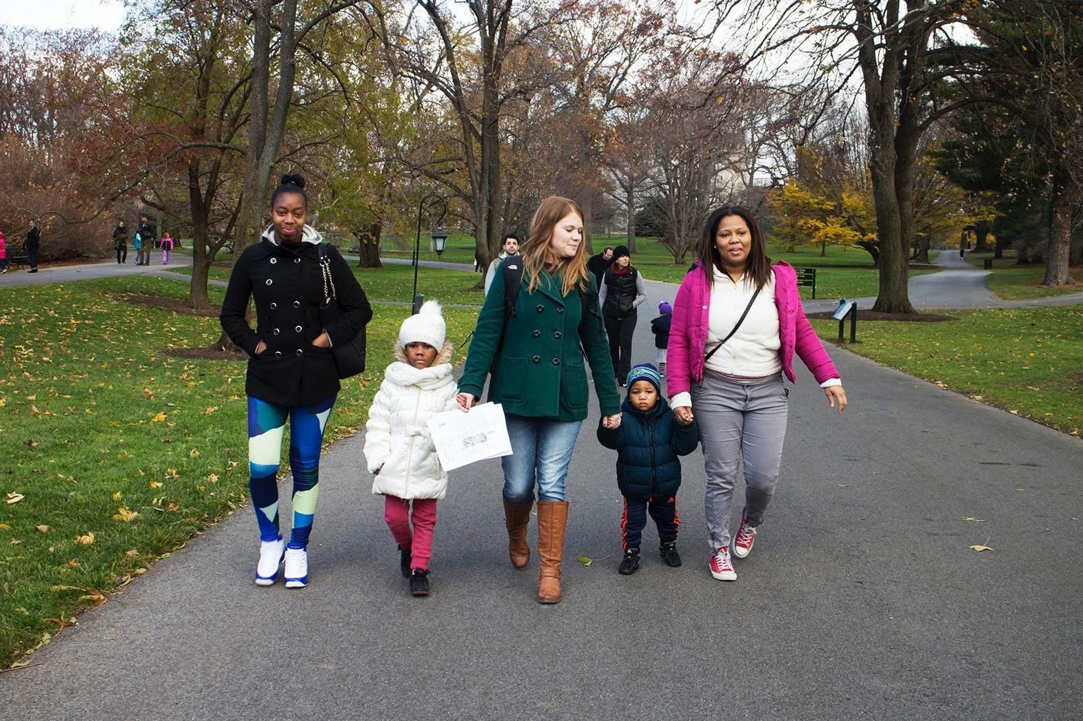 Group of five women and children walking in a park with autumn trees and paved paths.
