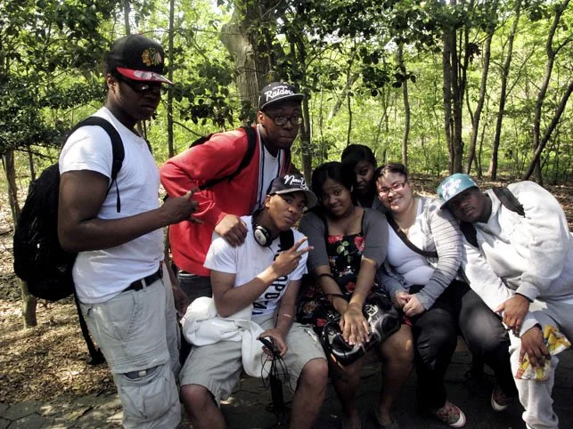 group of seven friends sitting and standing outdoors in a wooded area, some making peace signs, casually dressed.