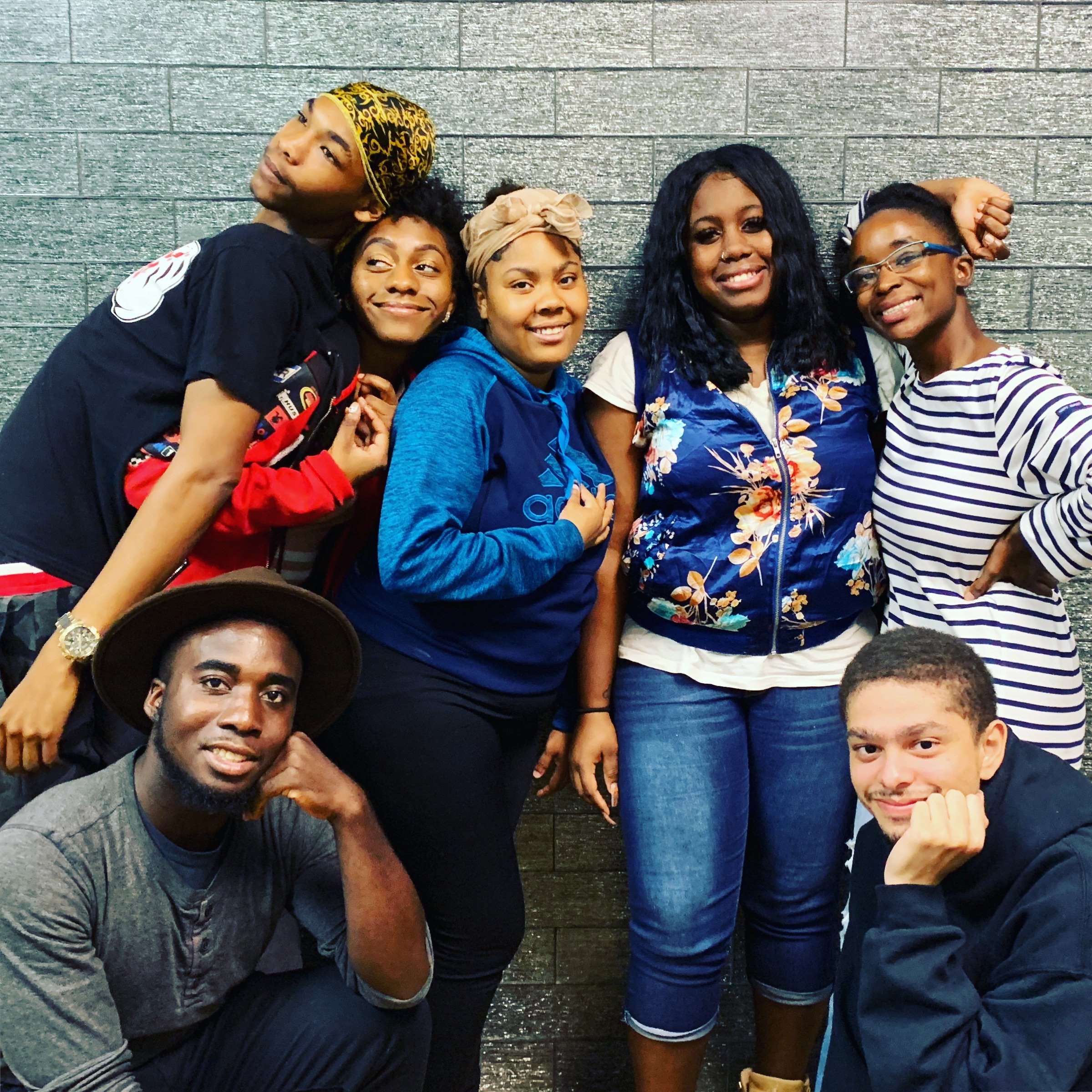 A group of seven diverse young adults posing together in front of a gray brick wall, some standing and some kneeling, smiling and making playful gestures.