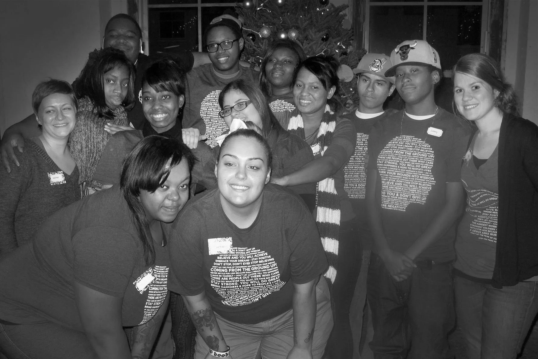 Group of people gathered in front of a Christmas tree, smiling for a photo, some wearing matching t-shirts.