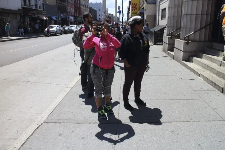 A group of five people standing in line on the sidewalk in an urban area, with some looking at their phones and one taking a photo.