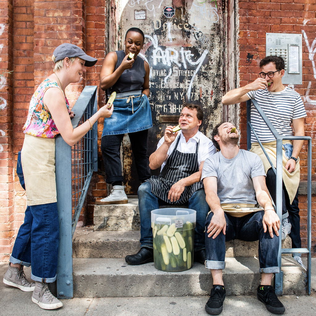 Group of five diverse friends, two women and three men, enjoying pickles and snacks on city stairs outside a brick building with graffiti, some wearing aprons and casual clothing.