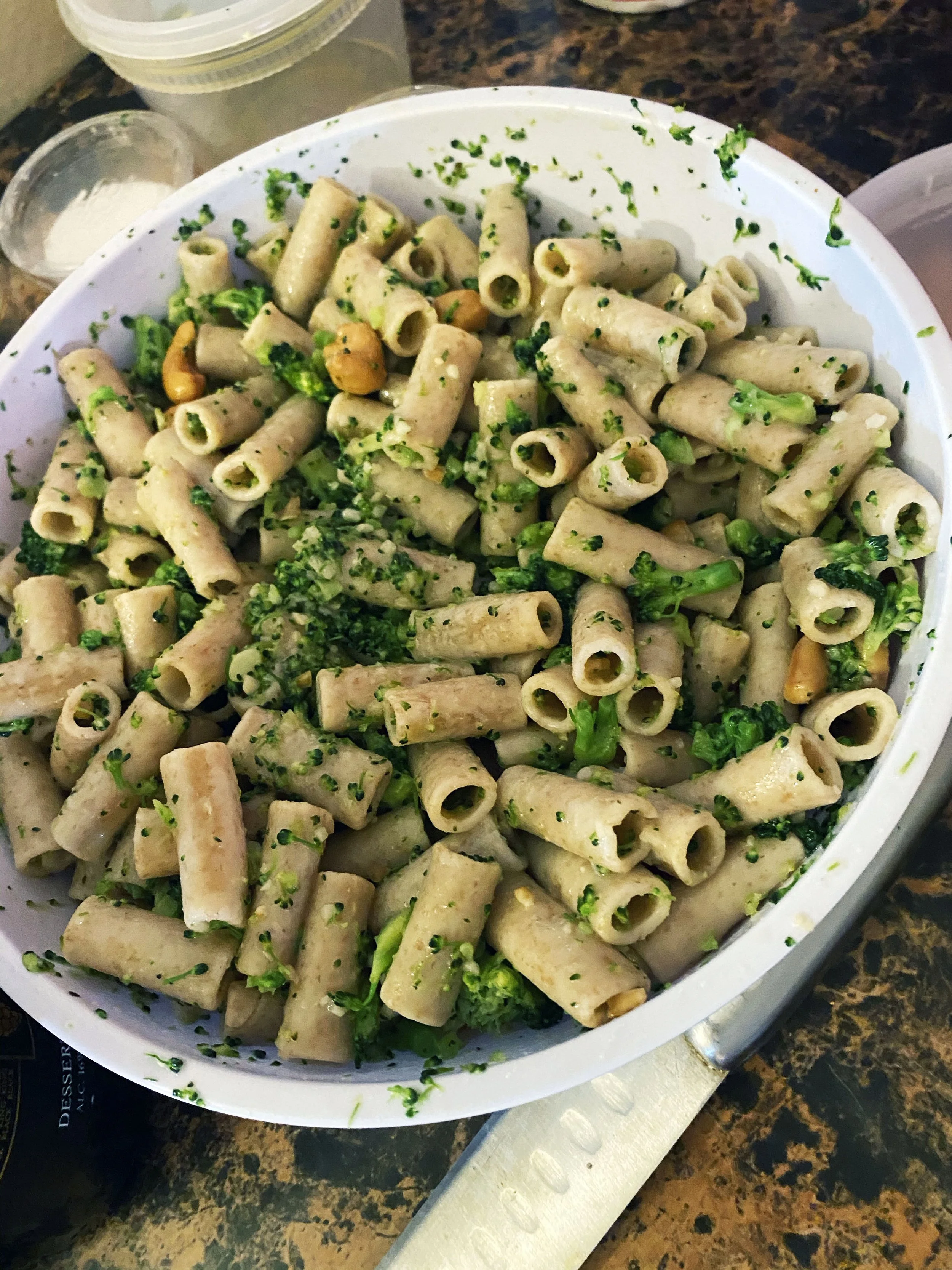 A large white bowl filled with pasta, broccoli, and chopped herbs, garnished with some peanuts, on a brown countertop with a knife nearby.