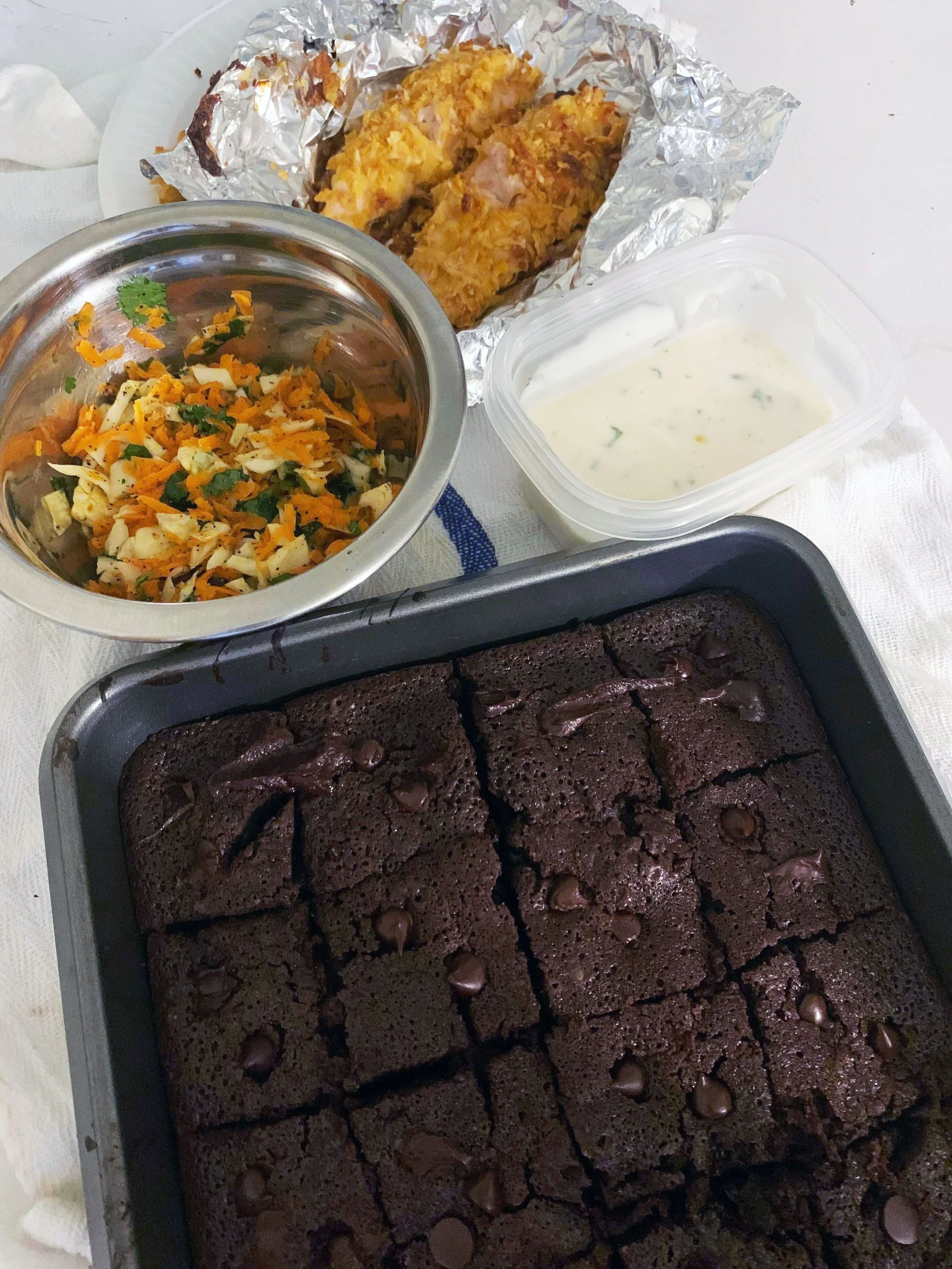 Baked chocolate brownies in a baking pan, a bowl of chopped vegetable salad, a container of ranch dressing, and a plate of fried chicken drumsticks with aluminum foil