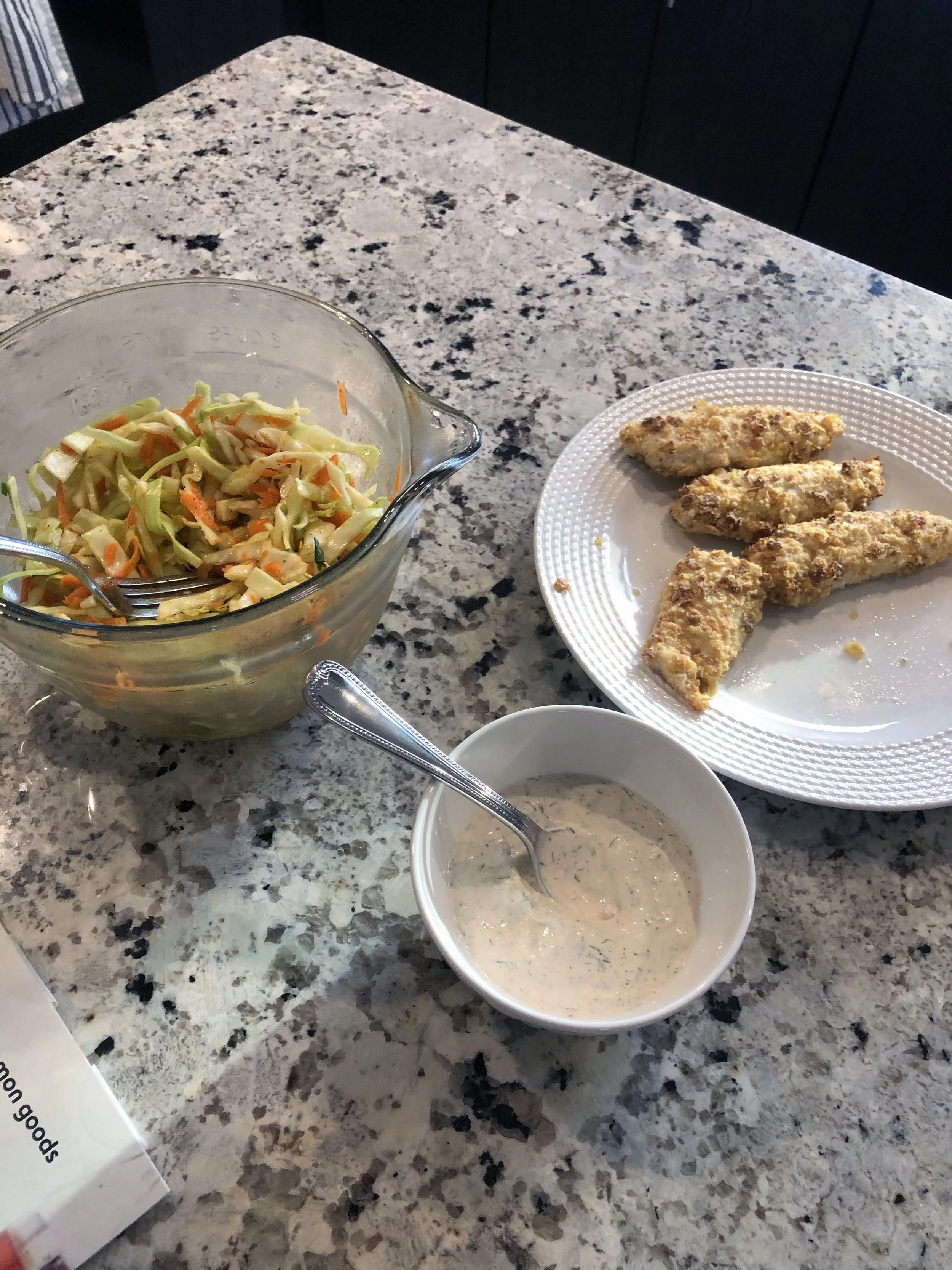 A glass bowl of coleslaw with shredded cabbage and carrots, a small bowl of creamy dressing, and a white plate with four pieces of fried chicken tenders on a speckled granite countertop.
