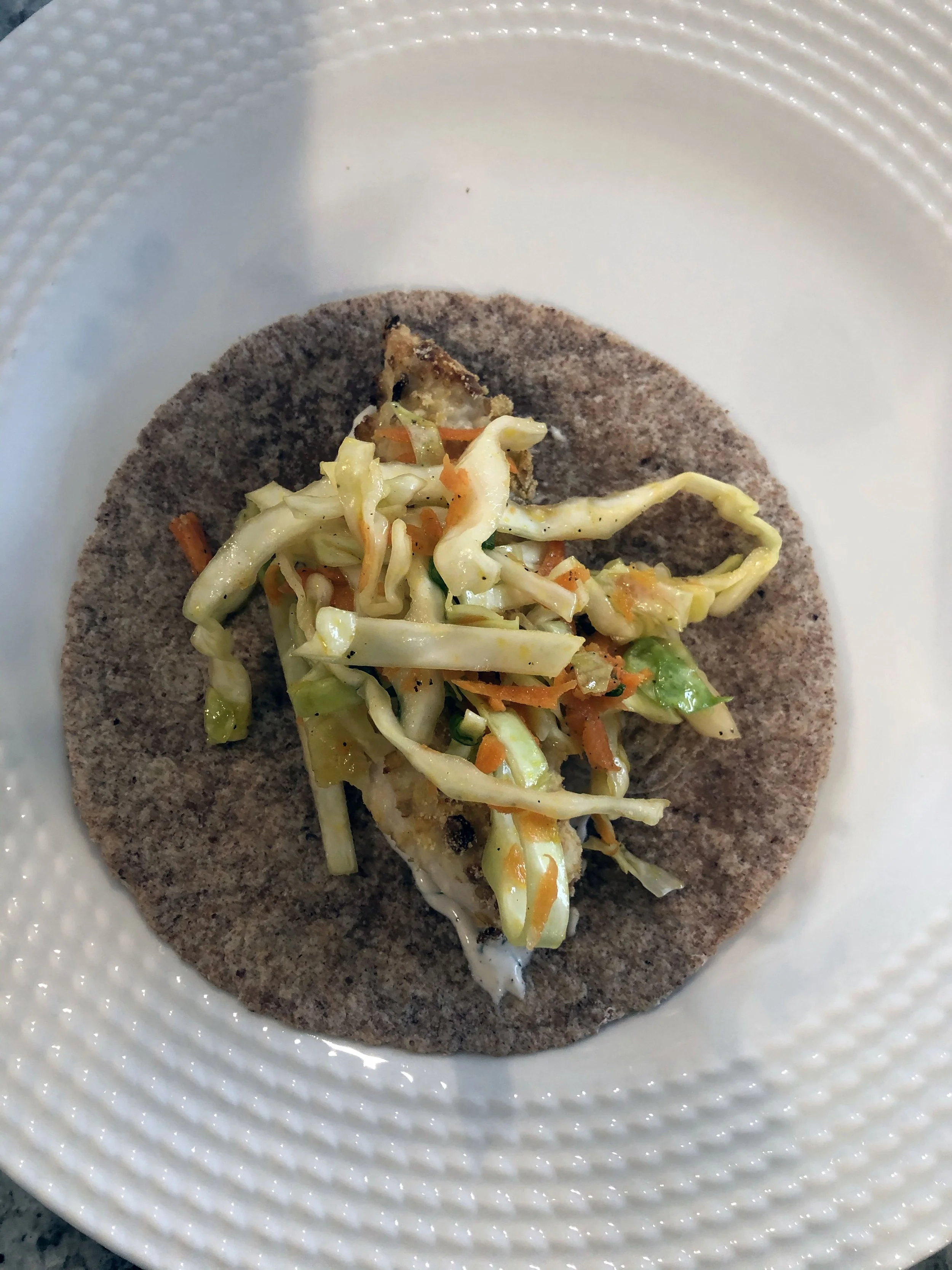 Close-up of an open-faced fish taco with shredded cabbage and carrots on a corn tortilla, on a textured white plate.