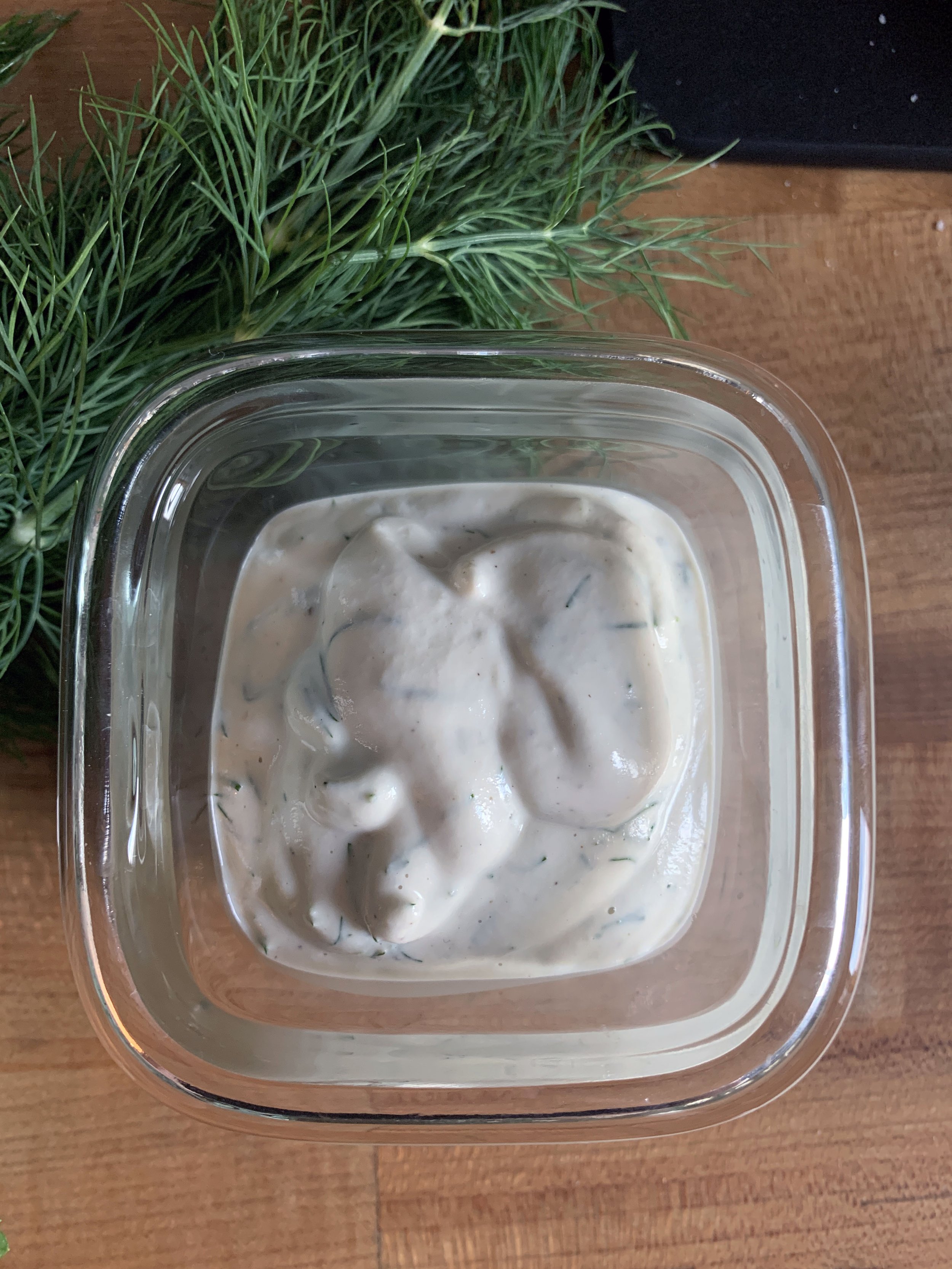 Creamy herb dip in a square glass container on a wooden surface with green herbs to the side.