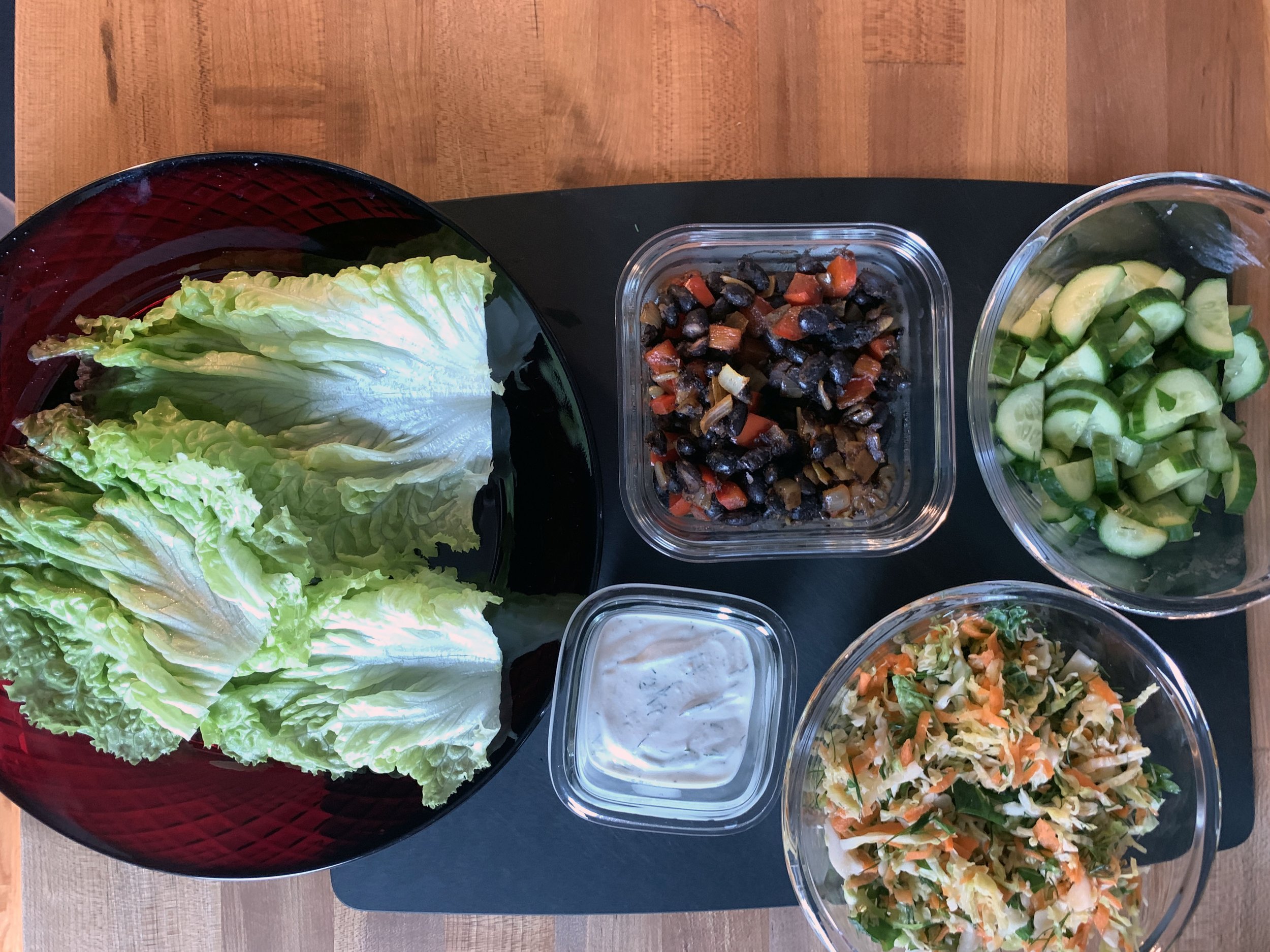 A variety of fresh vegetables and salad ingredients arranged on a black surface, including Romaine lettuce leaves in a red bowl, chopped cucumbers in a glass bowl, a mixture of chopped vegetables in a square glass container, a creamy dressing in a sm