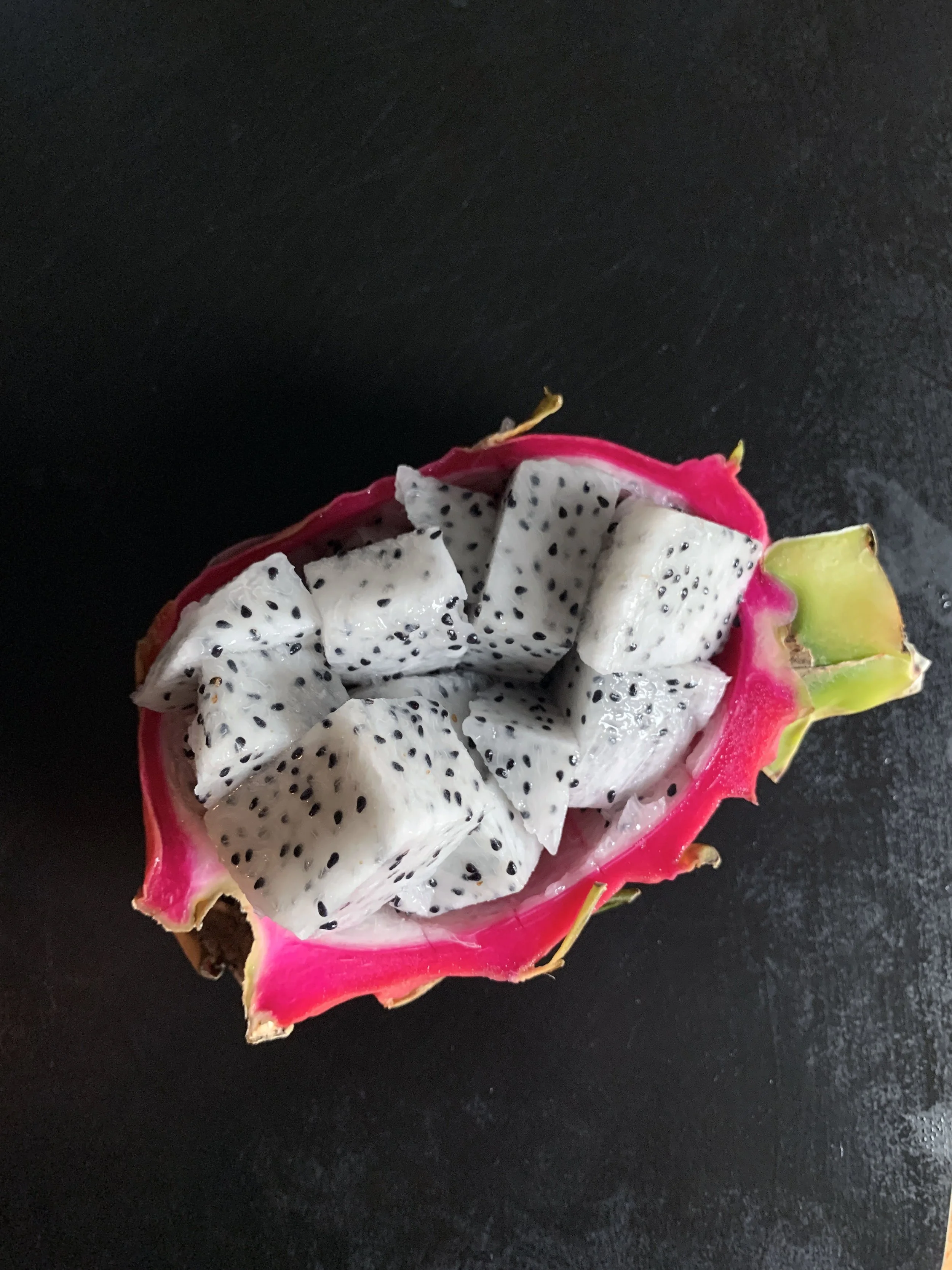 Half-cut dragon fruit with white flesh and black seeds filled with diced pieces of dragon fruit on a dark surface.