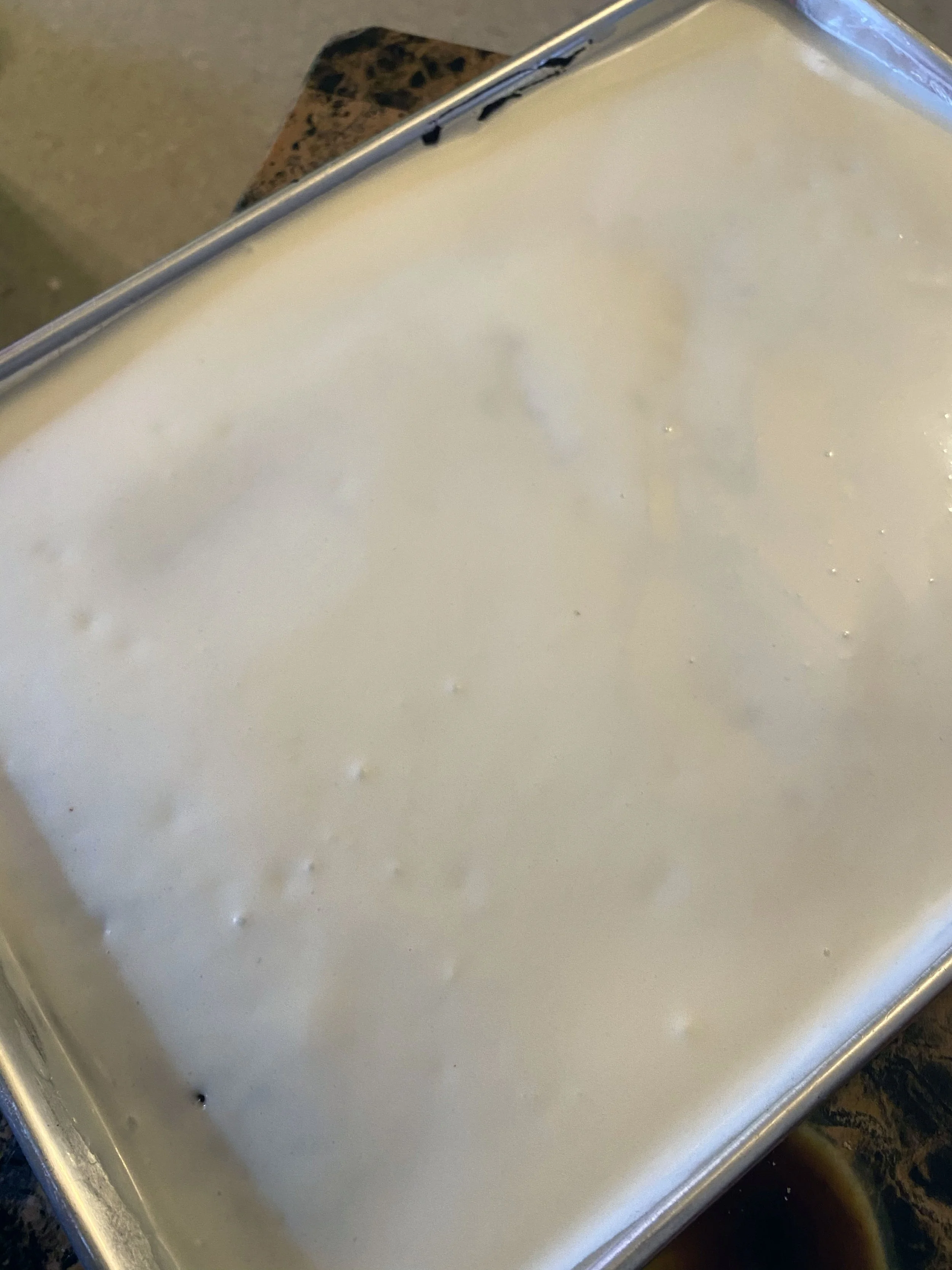A metal tray filled with a smooth, light-colored, creamy substance, possibly custard or pudding, placed on a speckled countertop.