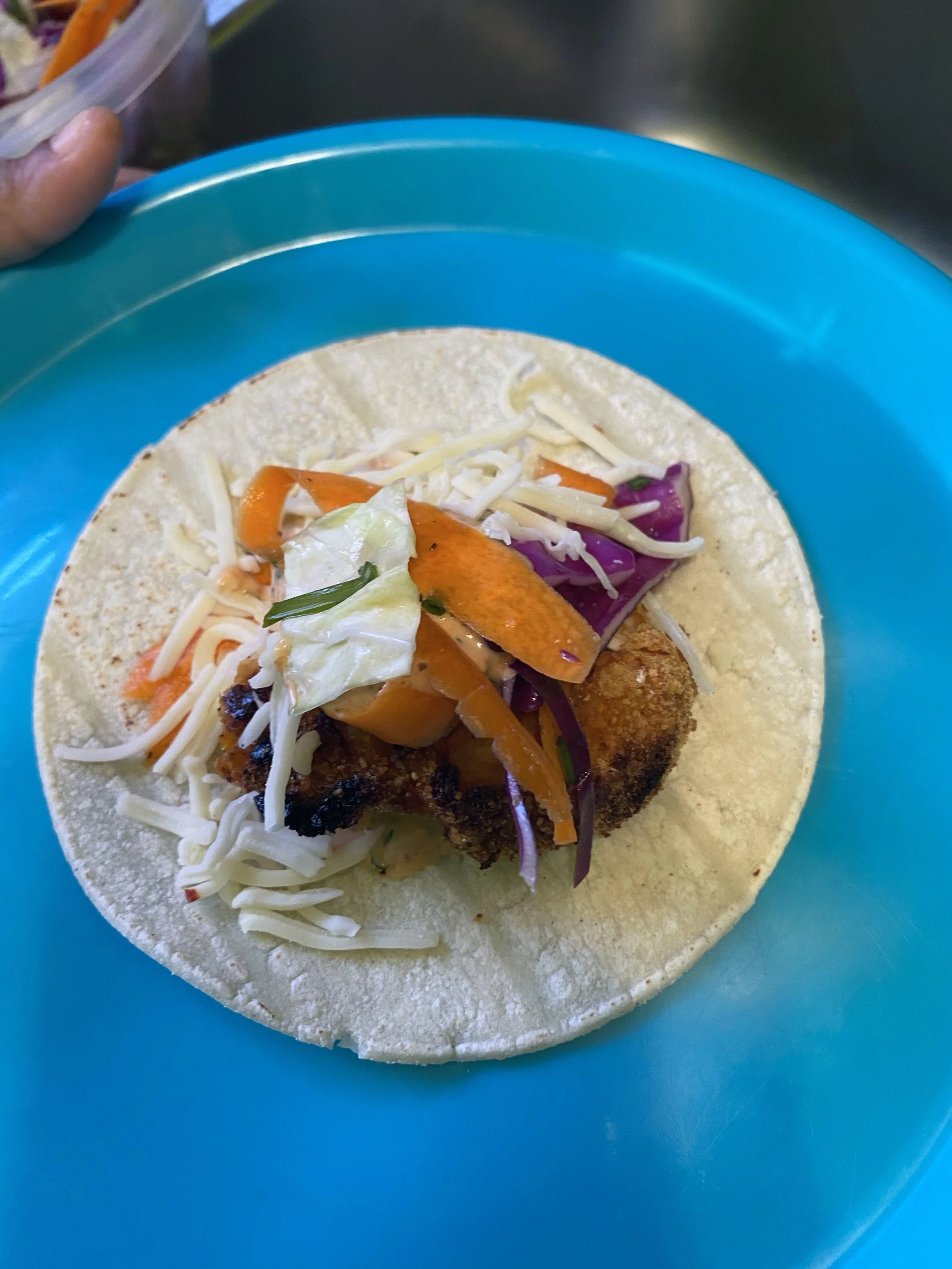 A taco with a soft flour tortilla, filled with fried fish, shredded cheese, lettuce, pickles, and shredded purple cabbage, served on a blue plate.