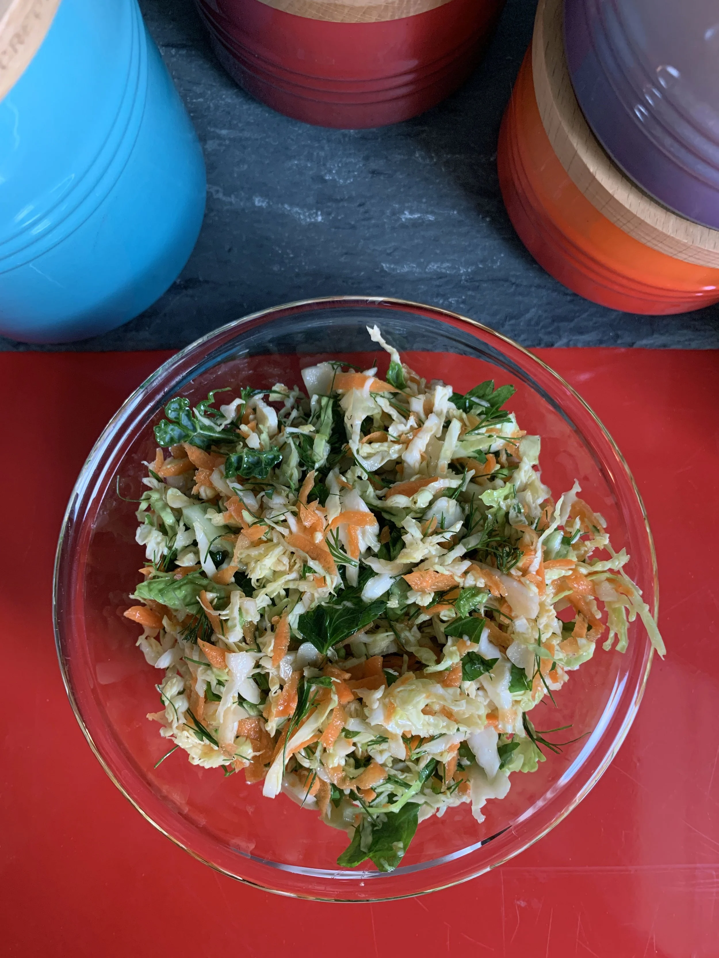 Bowl of shredded coleslaw salad with carrots and greens on a red surface. Several stacked colorful bowls are in the background.