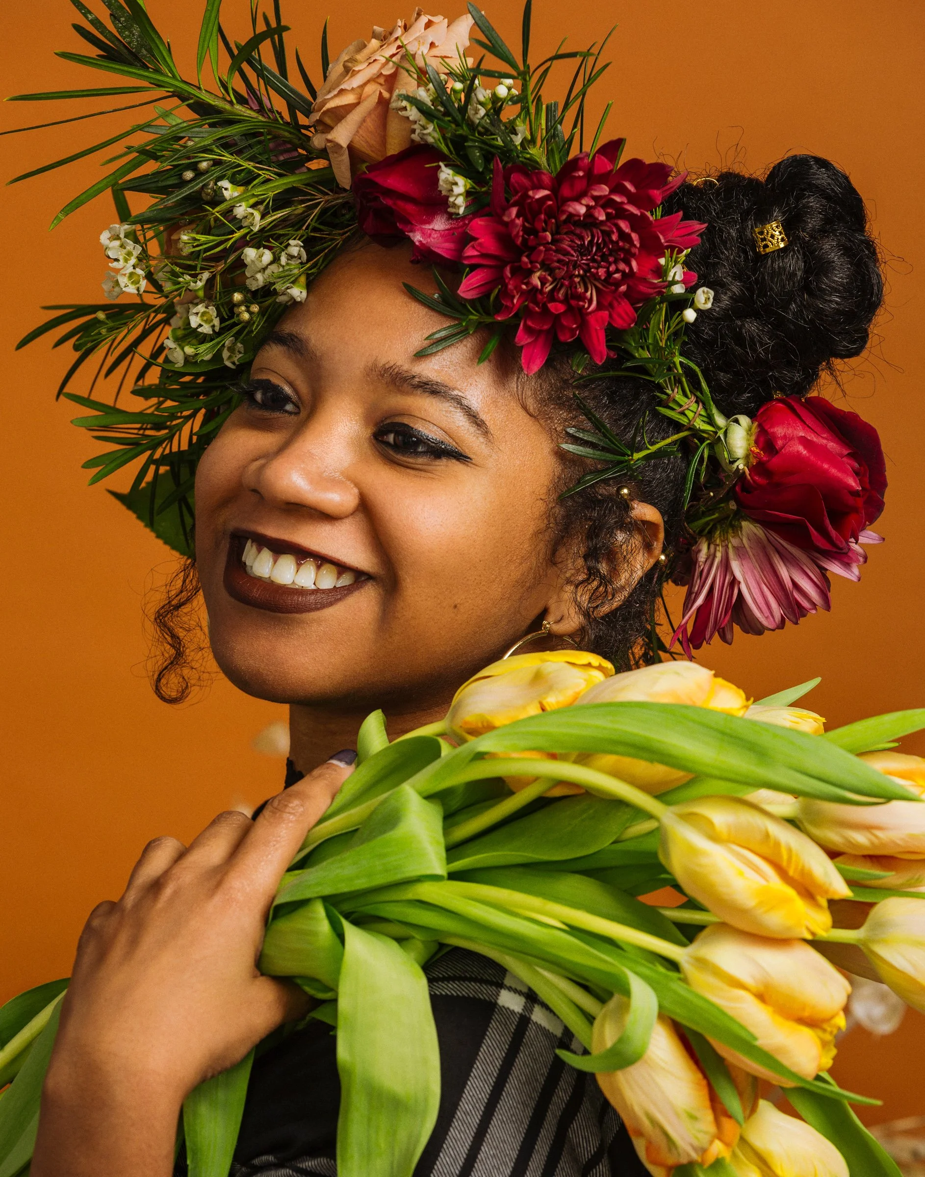 A smiling woman with a headpiece made of various colorful flowers and greenery, holding a large bouquet of yellow tulips against a warm orange background.