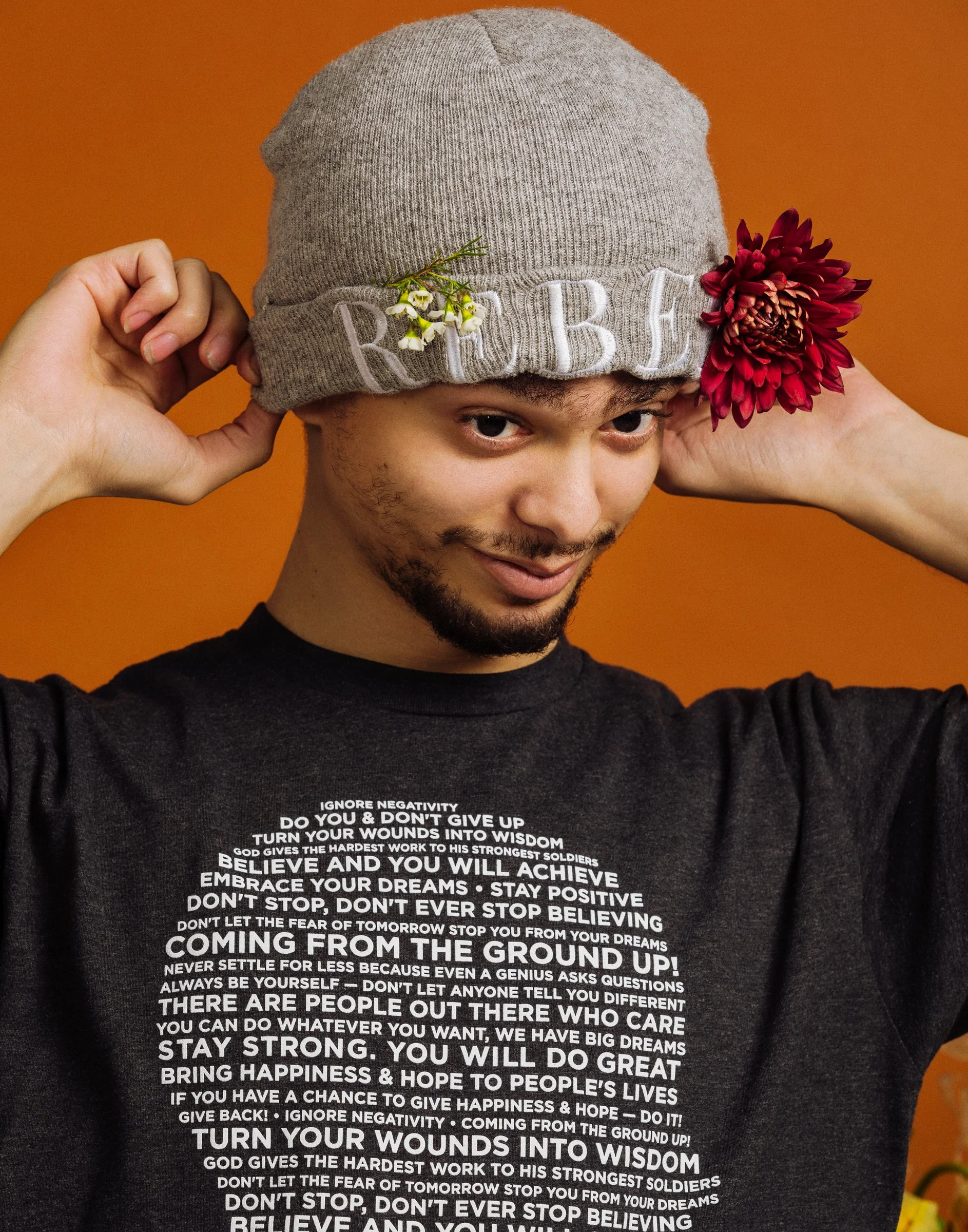 Young man wearing a gray beanie with flowers and the word 'REAL' embroidered on it, adjusting the beanie with both hands, against a warm orange background, wearing a black shirt with motivational text.