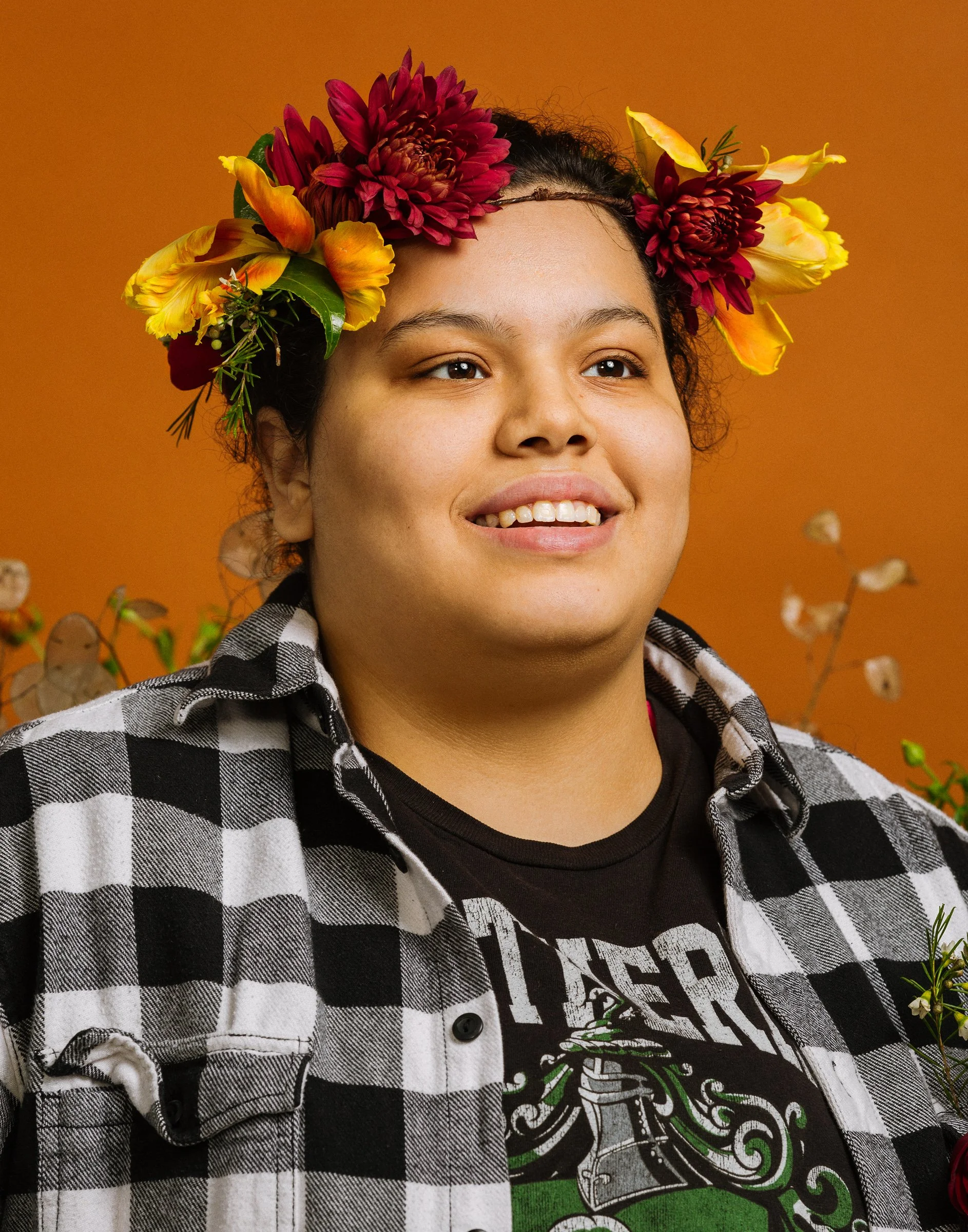 Person with a flower crown made of red and yellow flowers, smiling, wearing a black graphic t-shirt and a black-and-white checkered shirt, standing against an orange background with some plants visible.