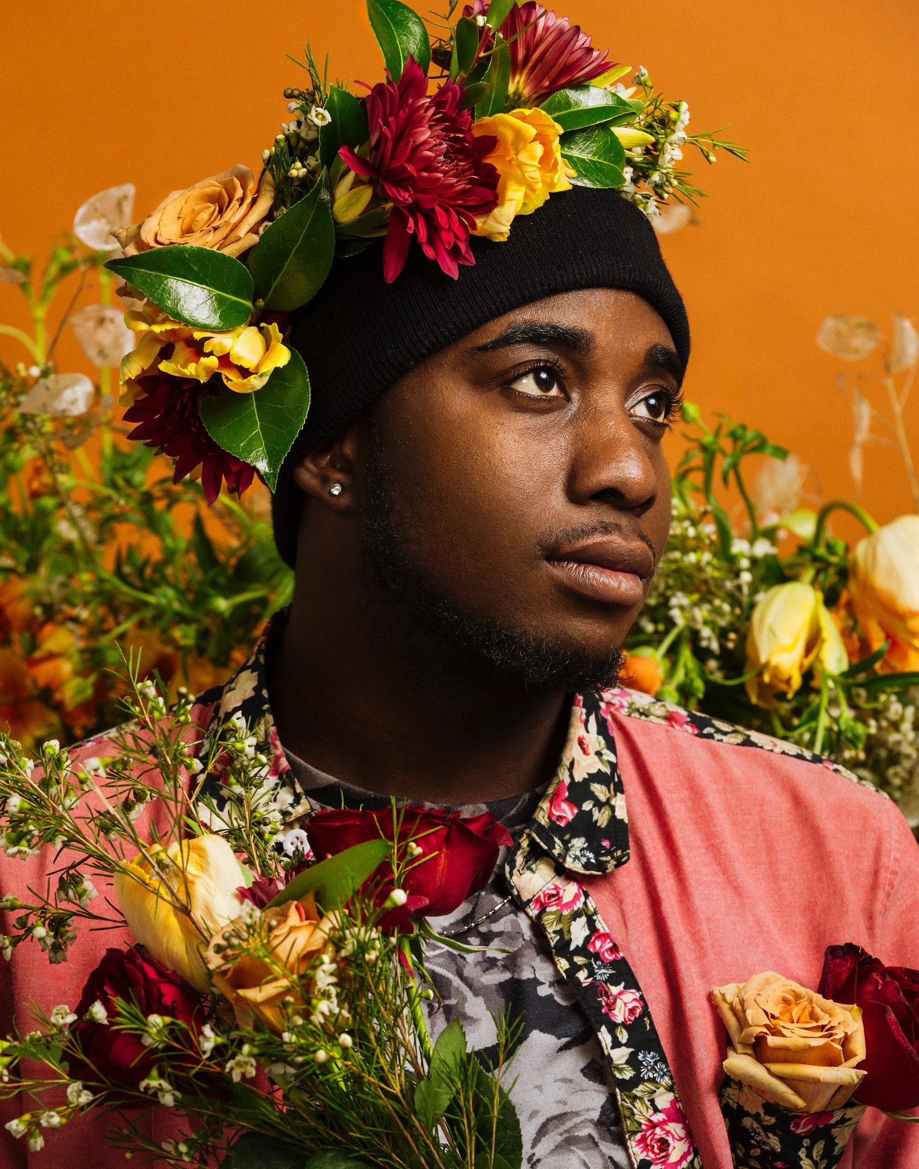 Young Black man with a black beanie decorated with colorful flowers, wearing a floral shirt and pink blazer, surrounded by an array of flowers against a warm orange background.