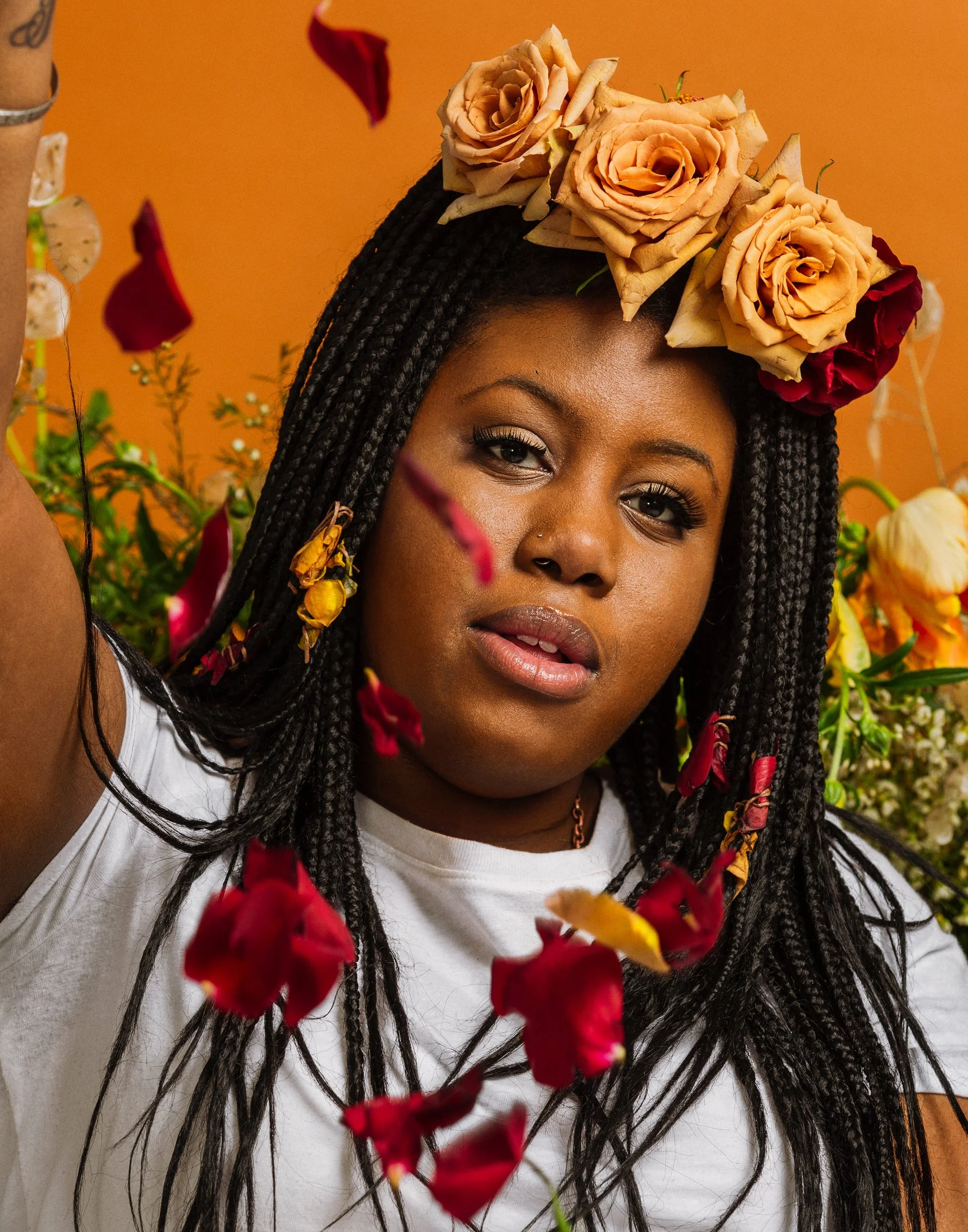 A woman with braided hair wearing a floral crown of roses, surrounded by flowers and rose petals, against an orange background.