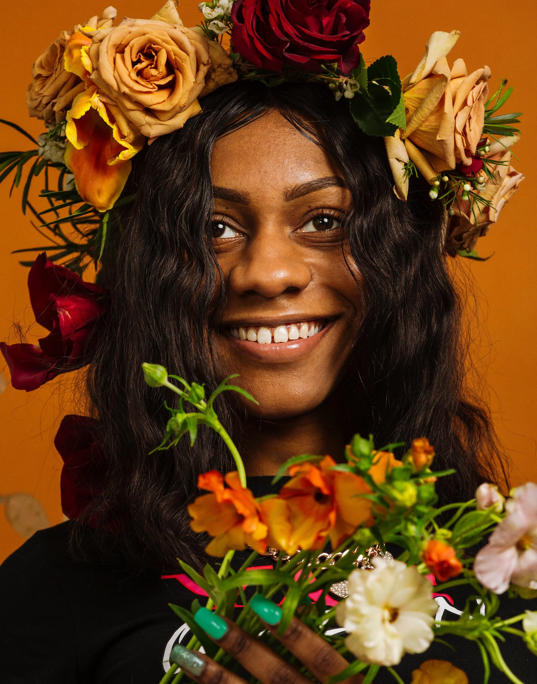 A woman with a big smile, wearing a floral crown made of various flowers, holds a bouquet of flowers, with an orange background.
