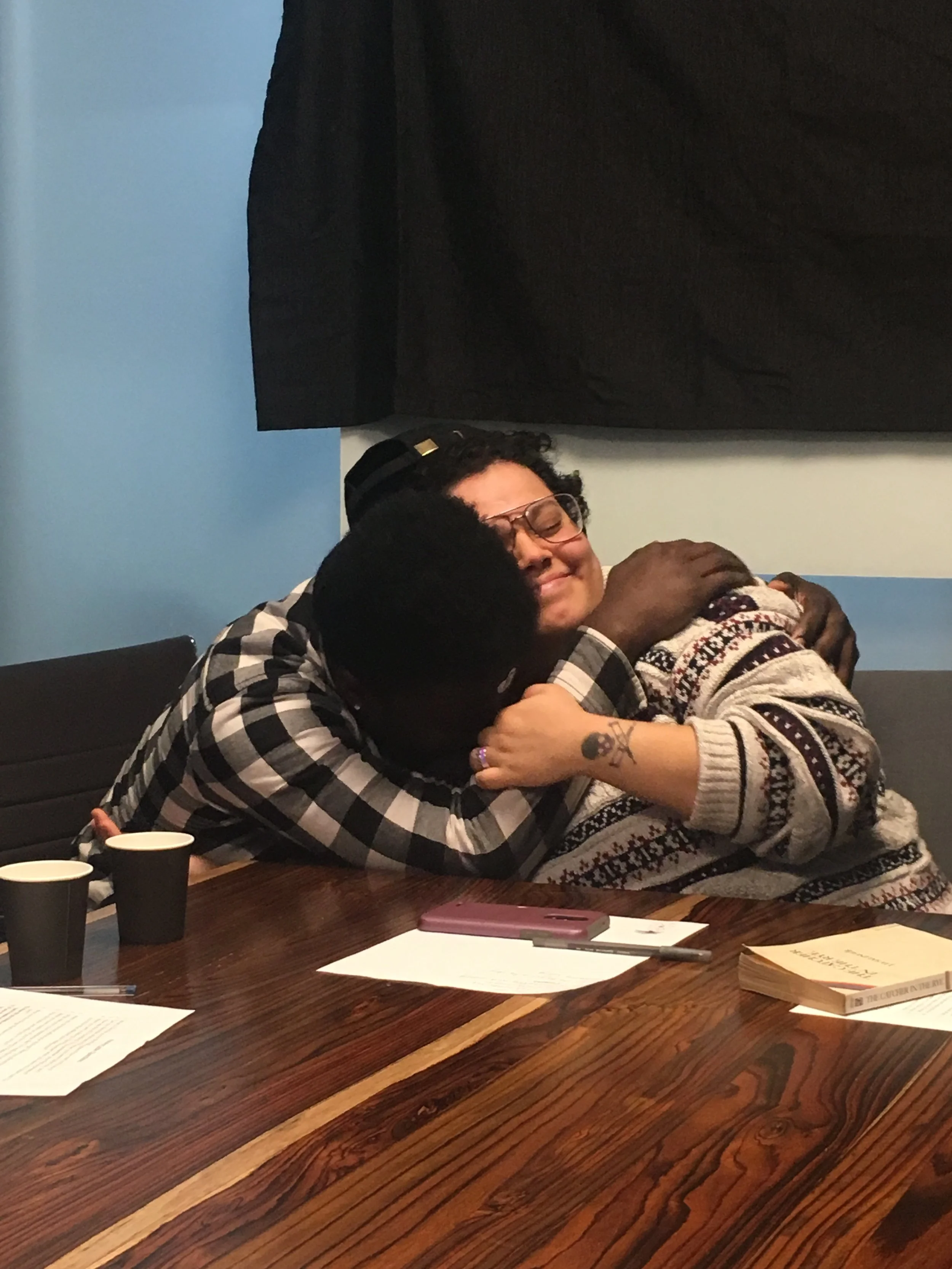 Two people hugging at a table, one with glasses and the other with a tattoo on her wrist, in a room with books and papers on the table.