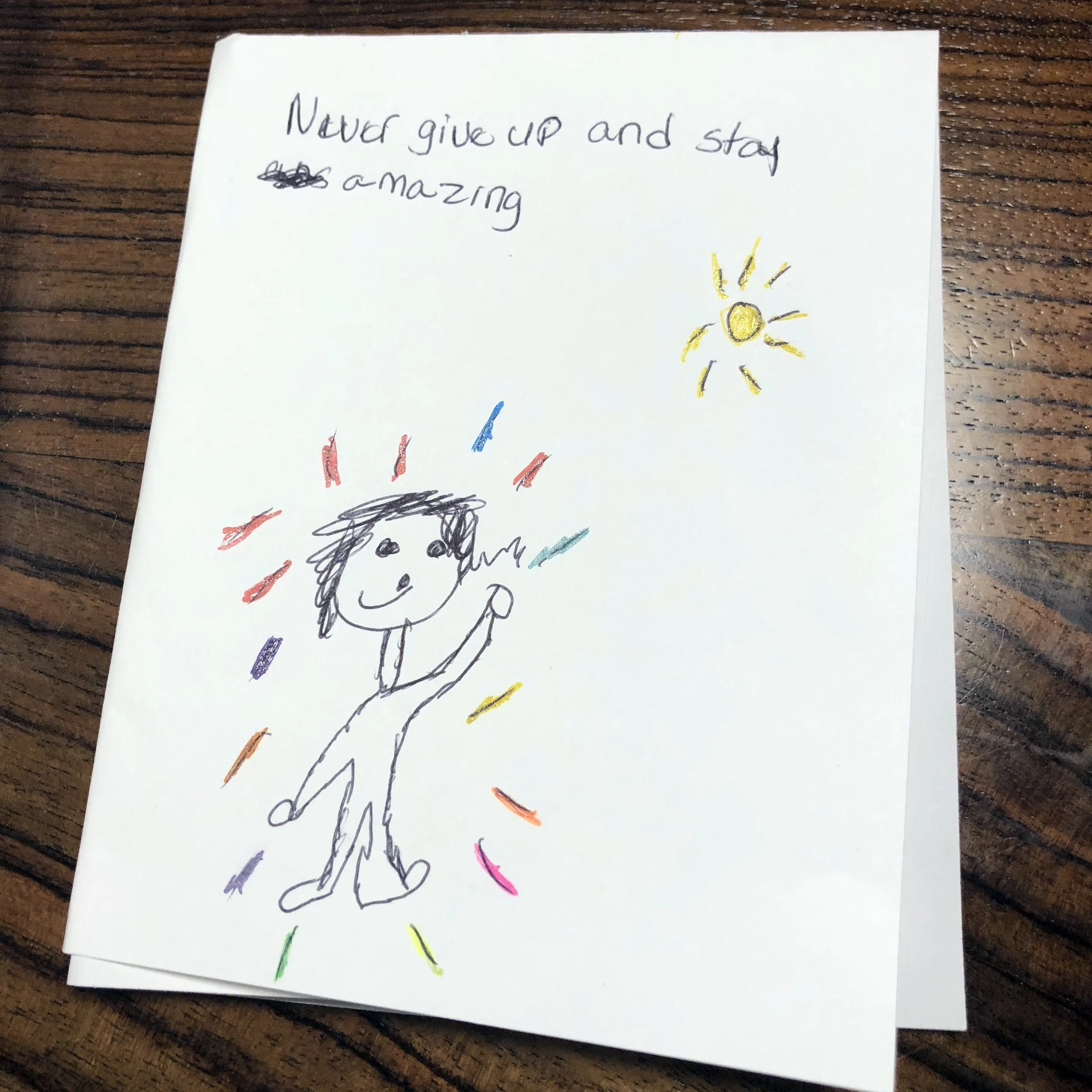 A child's drawing on a piece of white paper, depicting a person smiling and surrounded by colorful lines, with a yellow sun shining in the upper right corner. Handwritten text at the top says, 'Never give up and stay amazing'.