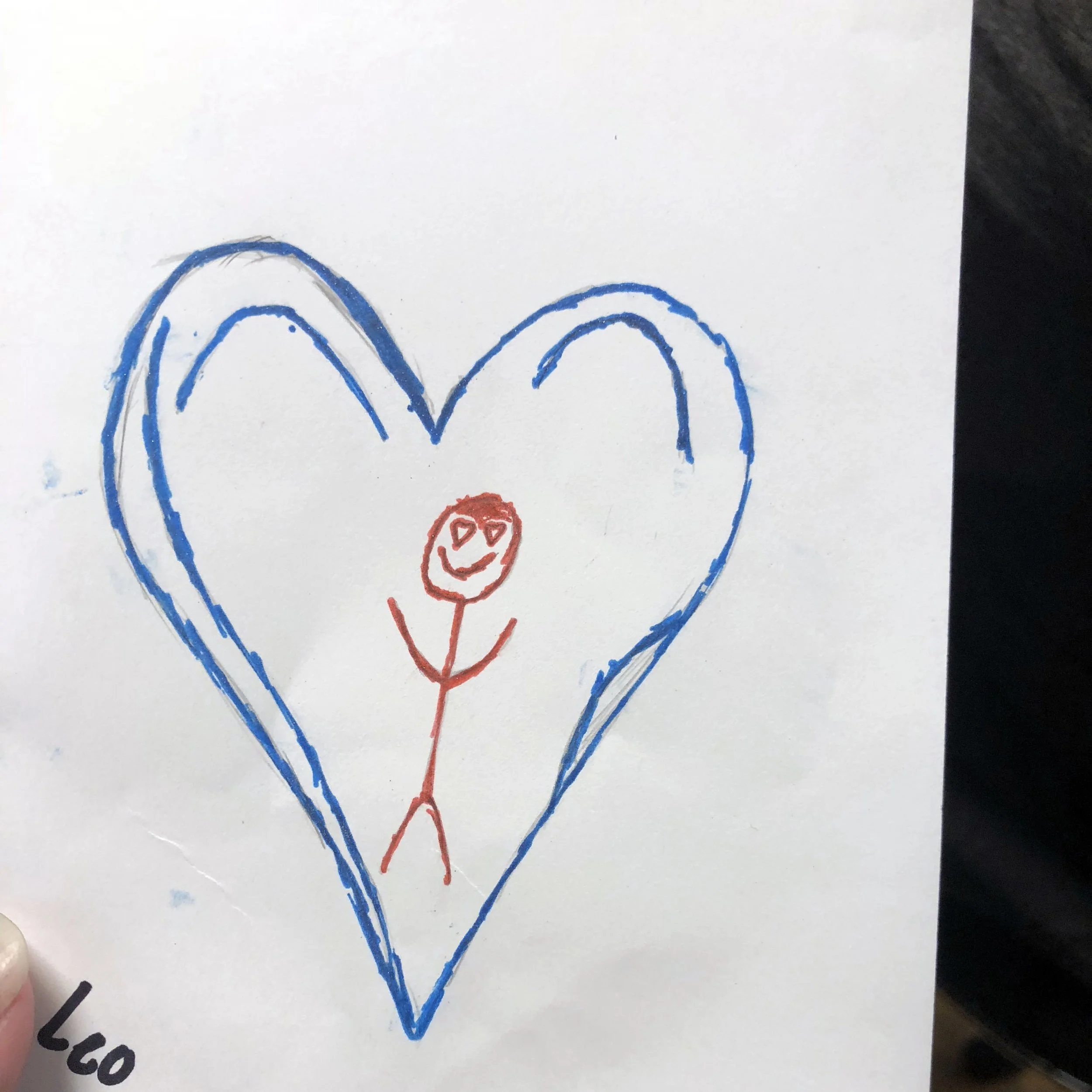 A child's drawing of a large blue heart with a smiling stick figure inside, drawn with red marker.