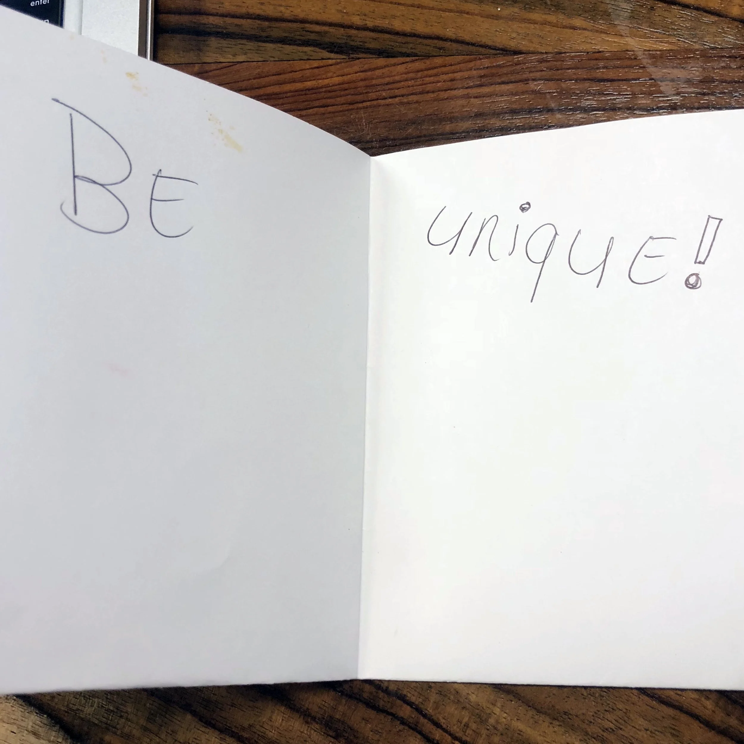 Open notebook with the words 'BE' and 'UNIQUE!' written in purple ink on white pages, placed on a wooden surface.