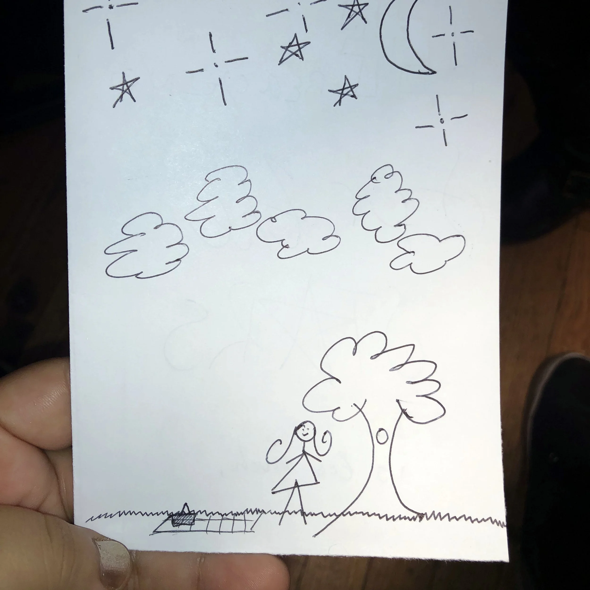 A hand-drawn sketch on white paper featuring a girl with pigtails standing near a tree with a swing, several clouds, stars, and a crescent moon in the night sky.