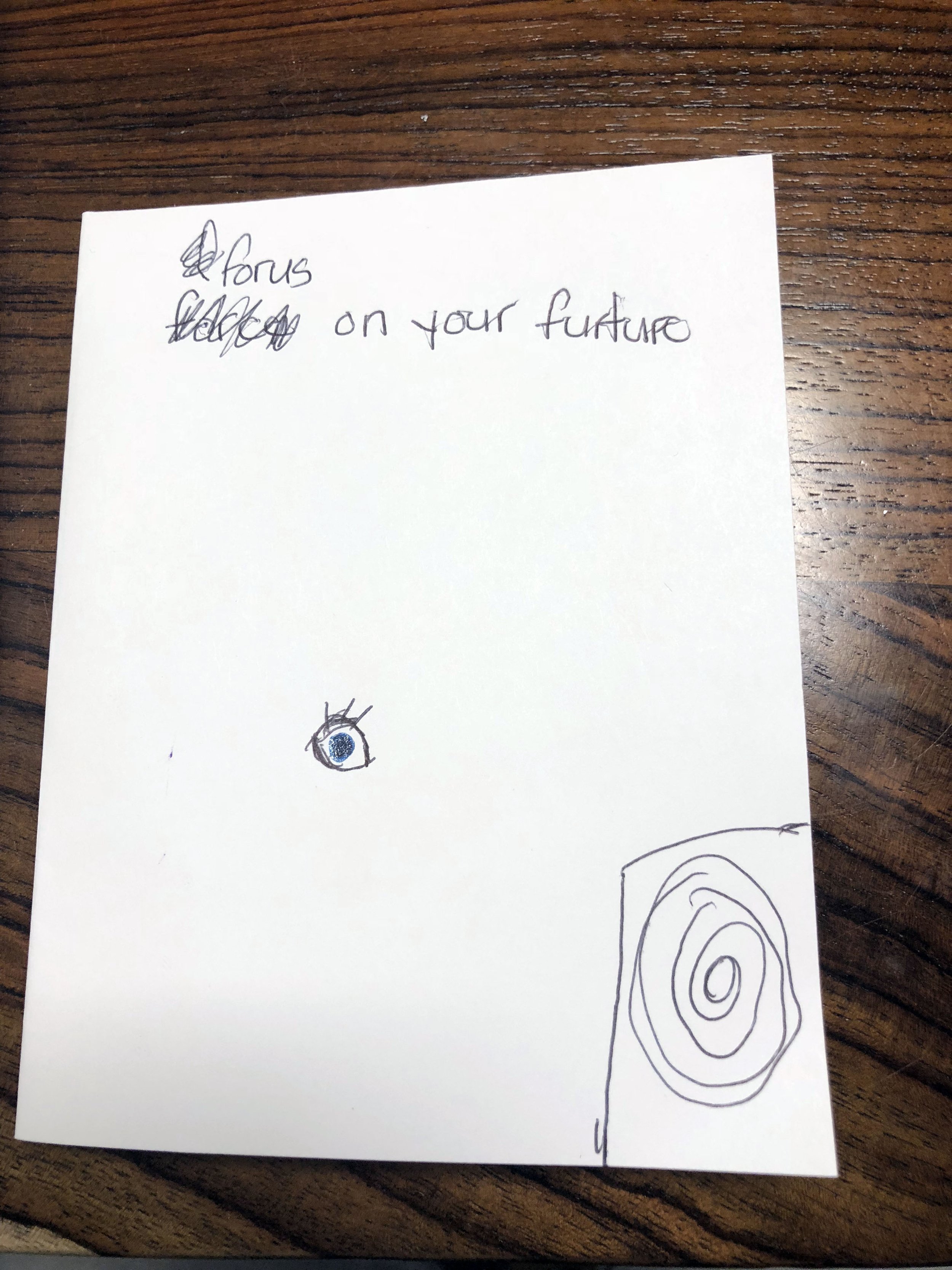 A handwritten note on a white paper with a drawing of an eye and a spiral pattern, placed on a wooden surface.
